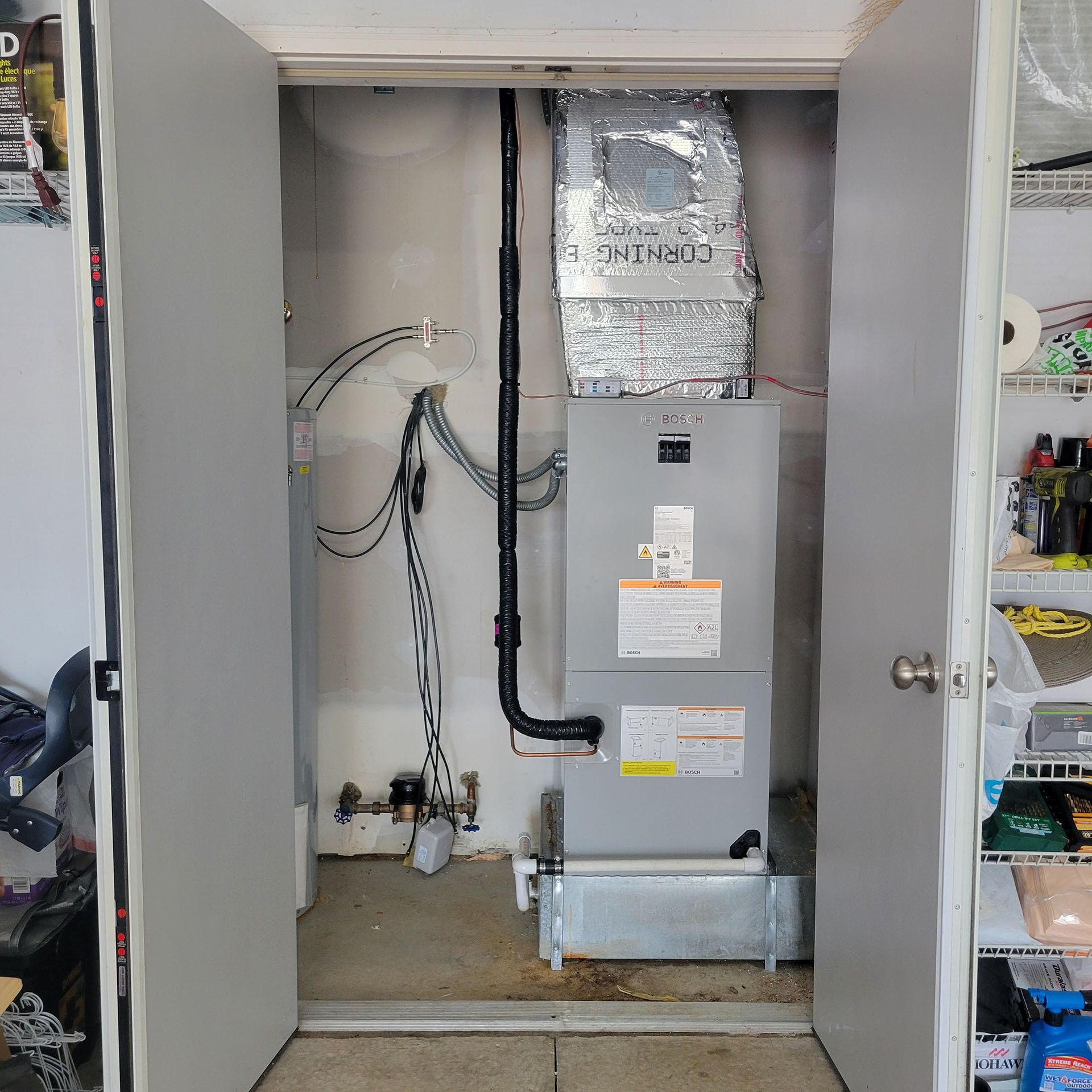 HVAC unit inside a closet, framed by open grey doors.  Piping, wiring, and shelving are also visible.
