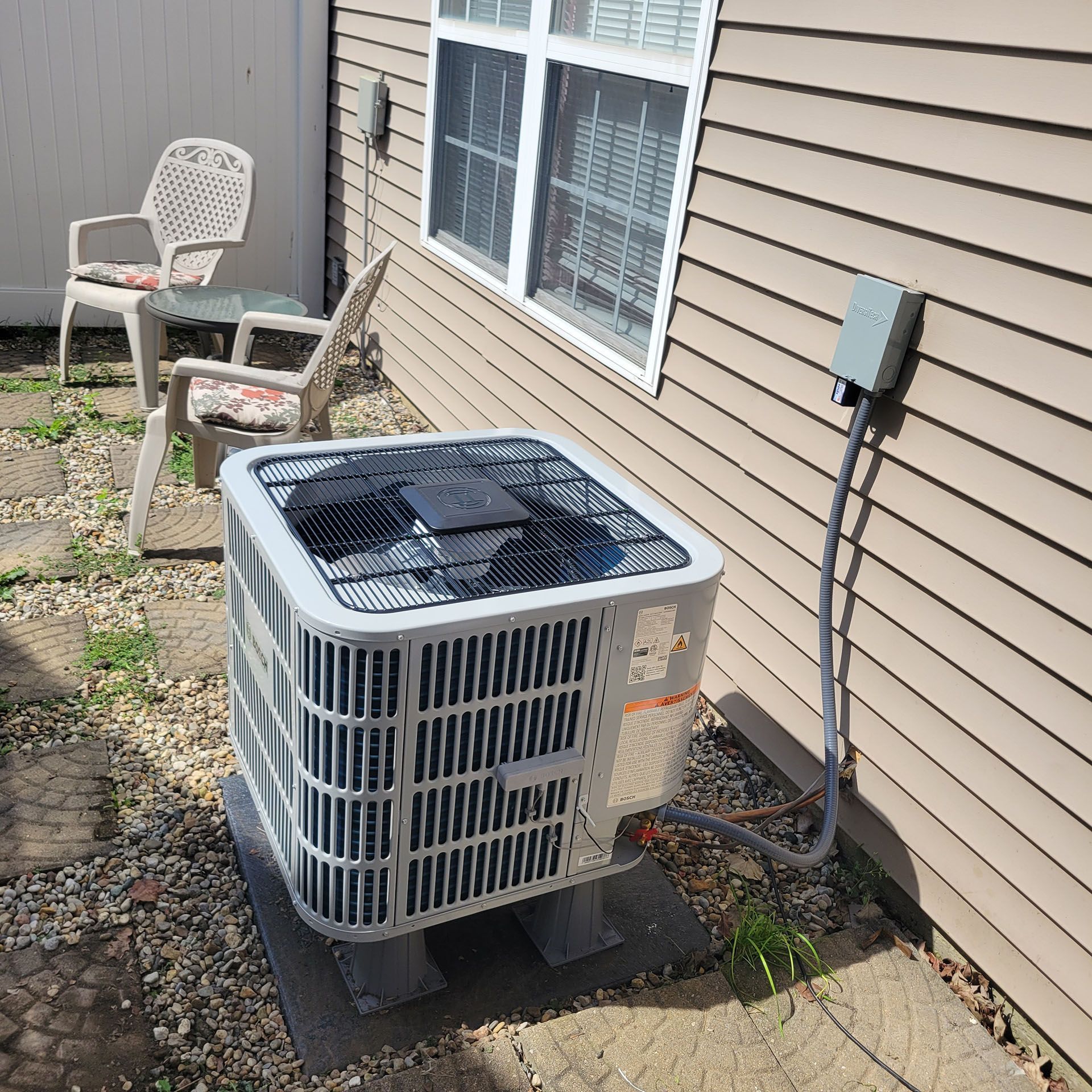 Air conditioner unit outside a building, near a window and chairs on a stone patio.