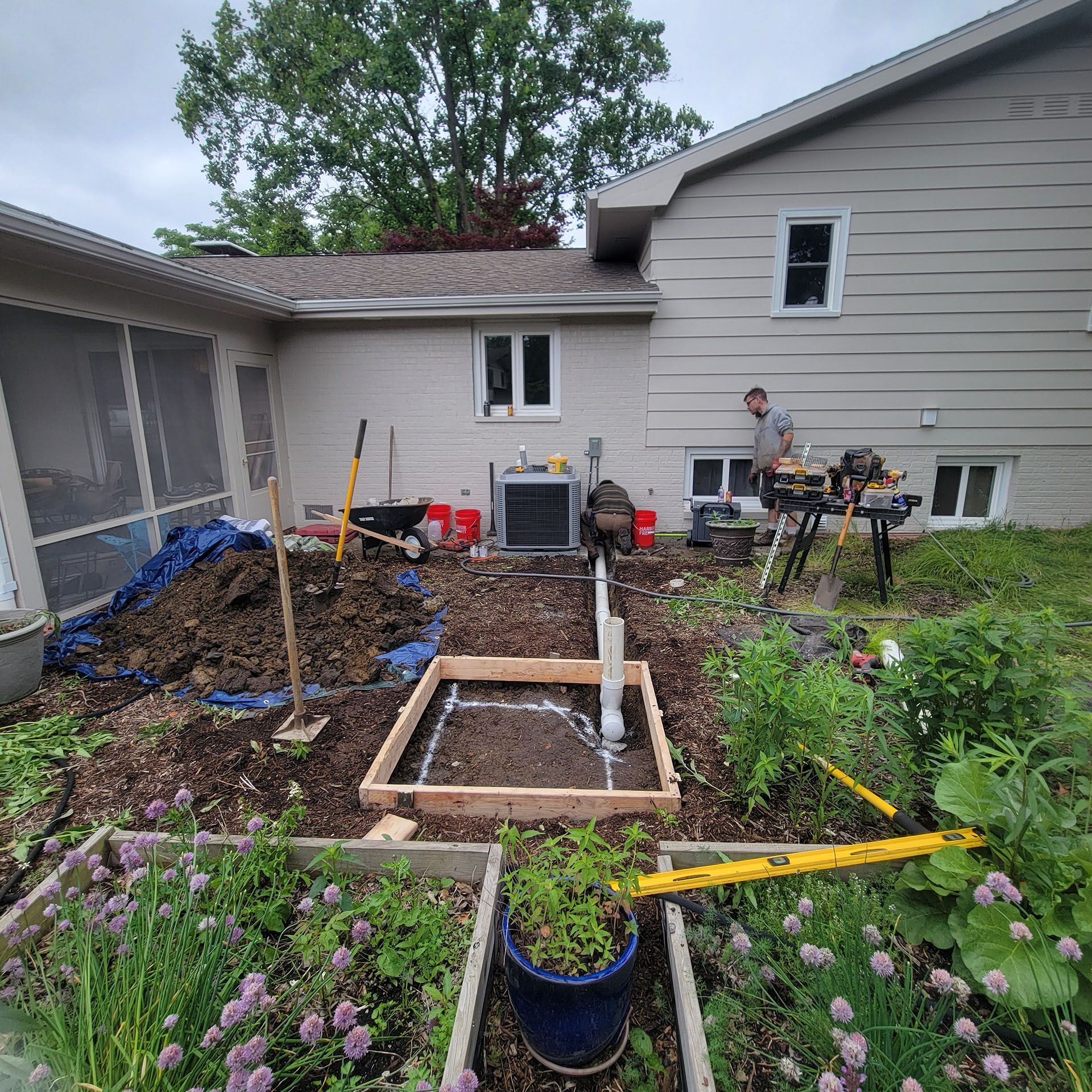 Construction site with a house in the background. A man works near a table, and a wooden frame sits on the ground.