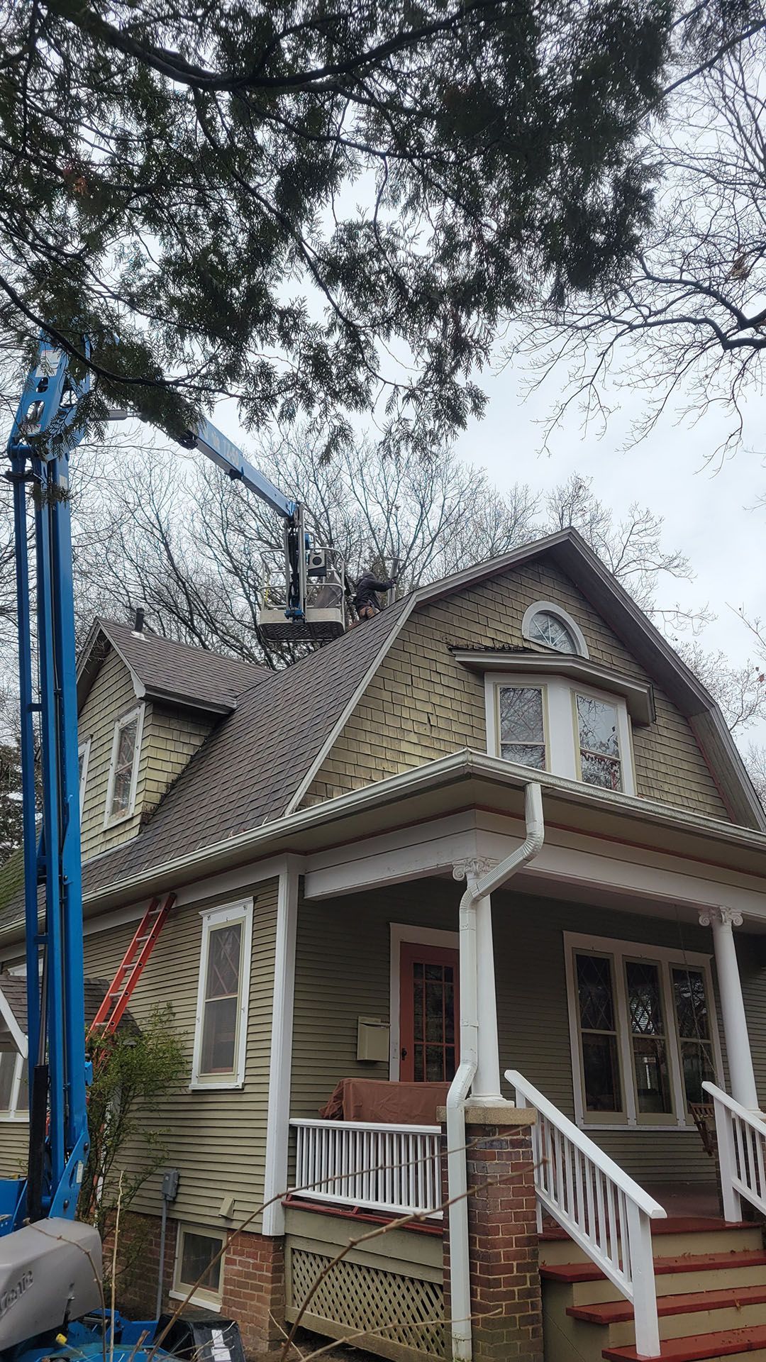 A blue lift platform reaches a house roof, likely for repair. The house is green, with white trim and a porch.