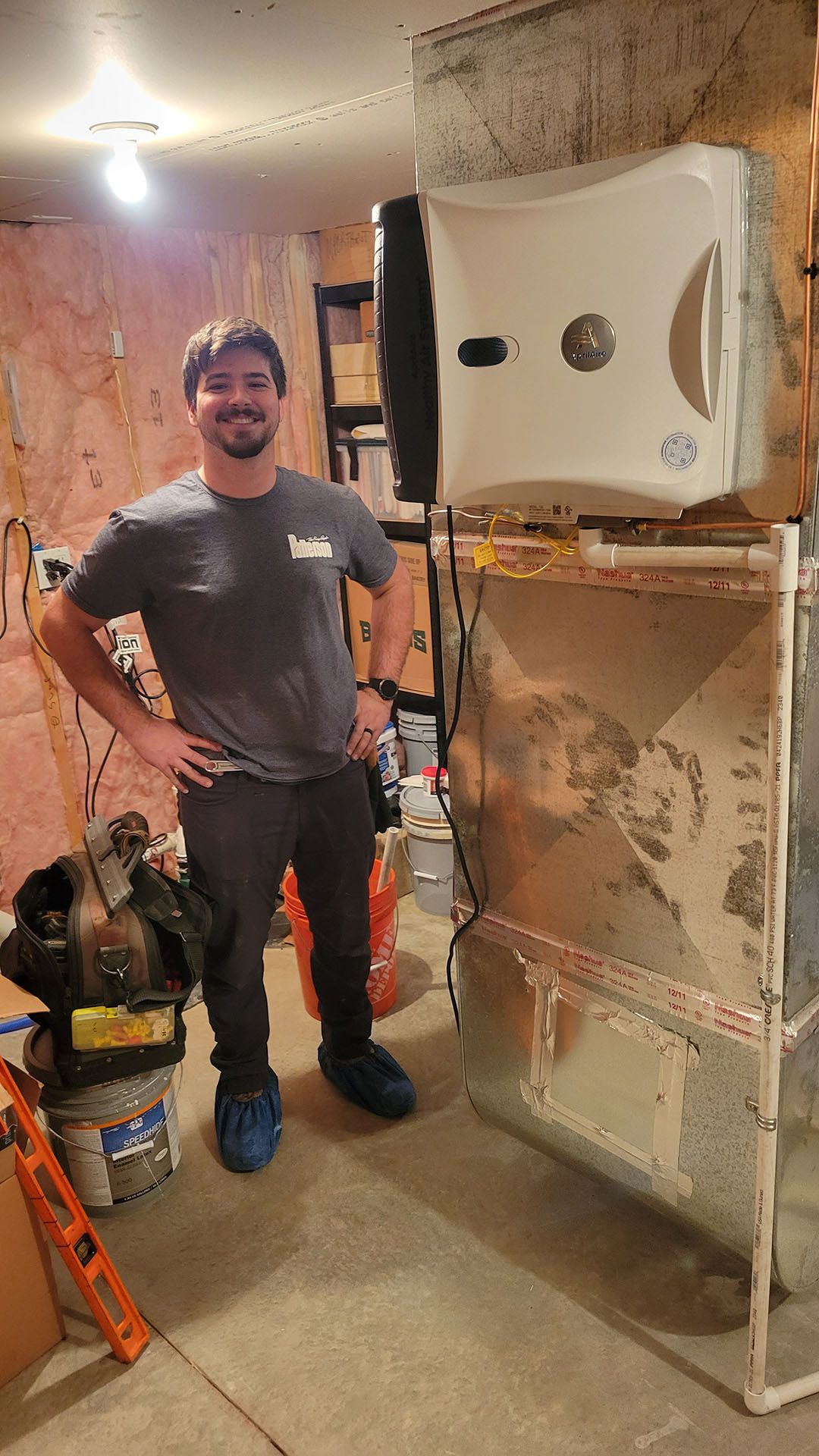 Man stands by HVAC unit, tools nearby. He wears a grey shirt, black pants, and blue shoes, smiling in a basement.