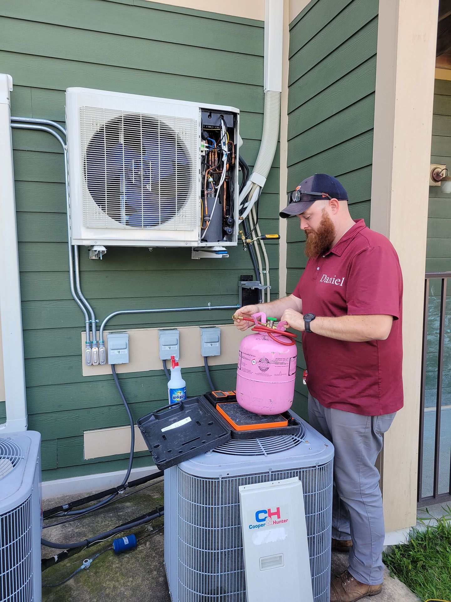 HVAC technician near outdoor AC unit and open wall-mounted air handler, handling pink refrigerant tank.