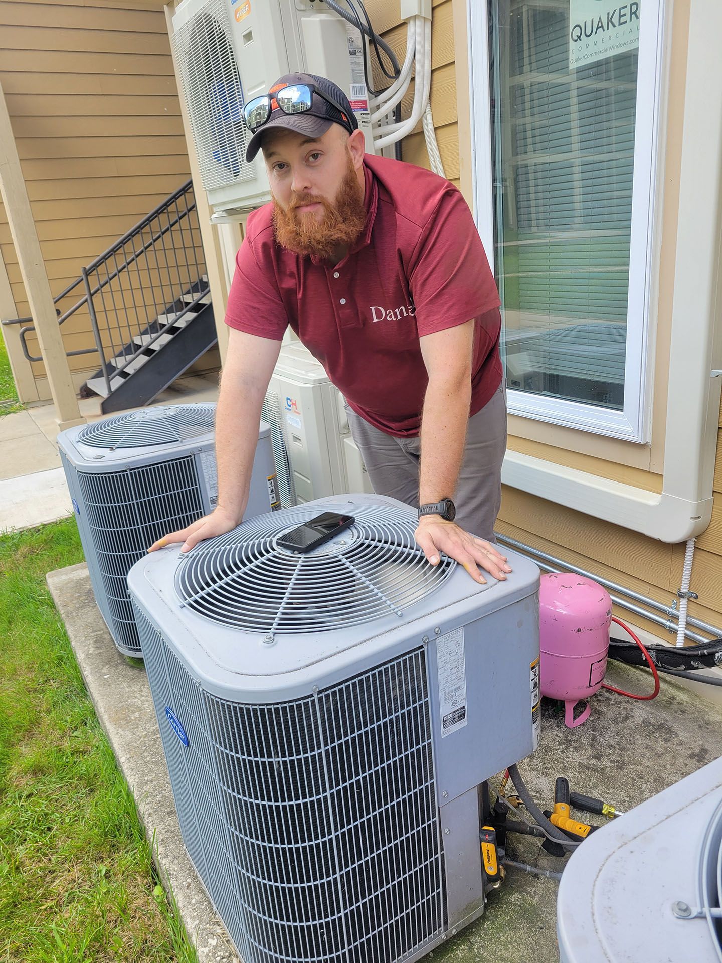 Man with beard, wearing red polo shirt, leaning over AC unit outside.  Building background.