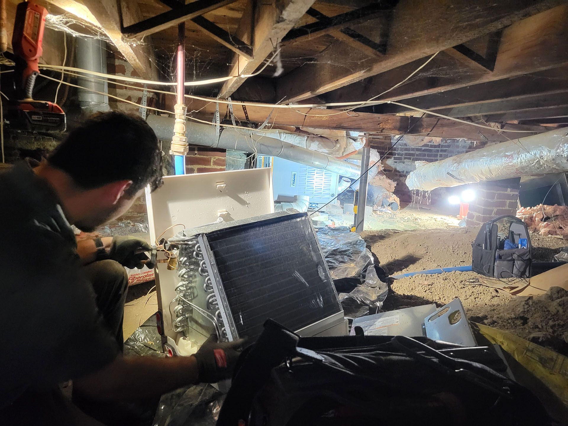 A person working on an air conditioning unit in a crawl space. Low lighting, visible insulation, and equipment.
