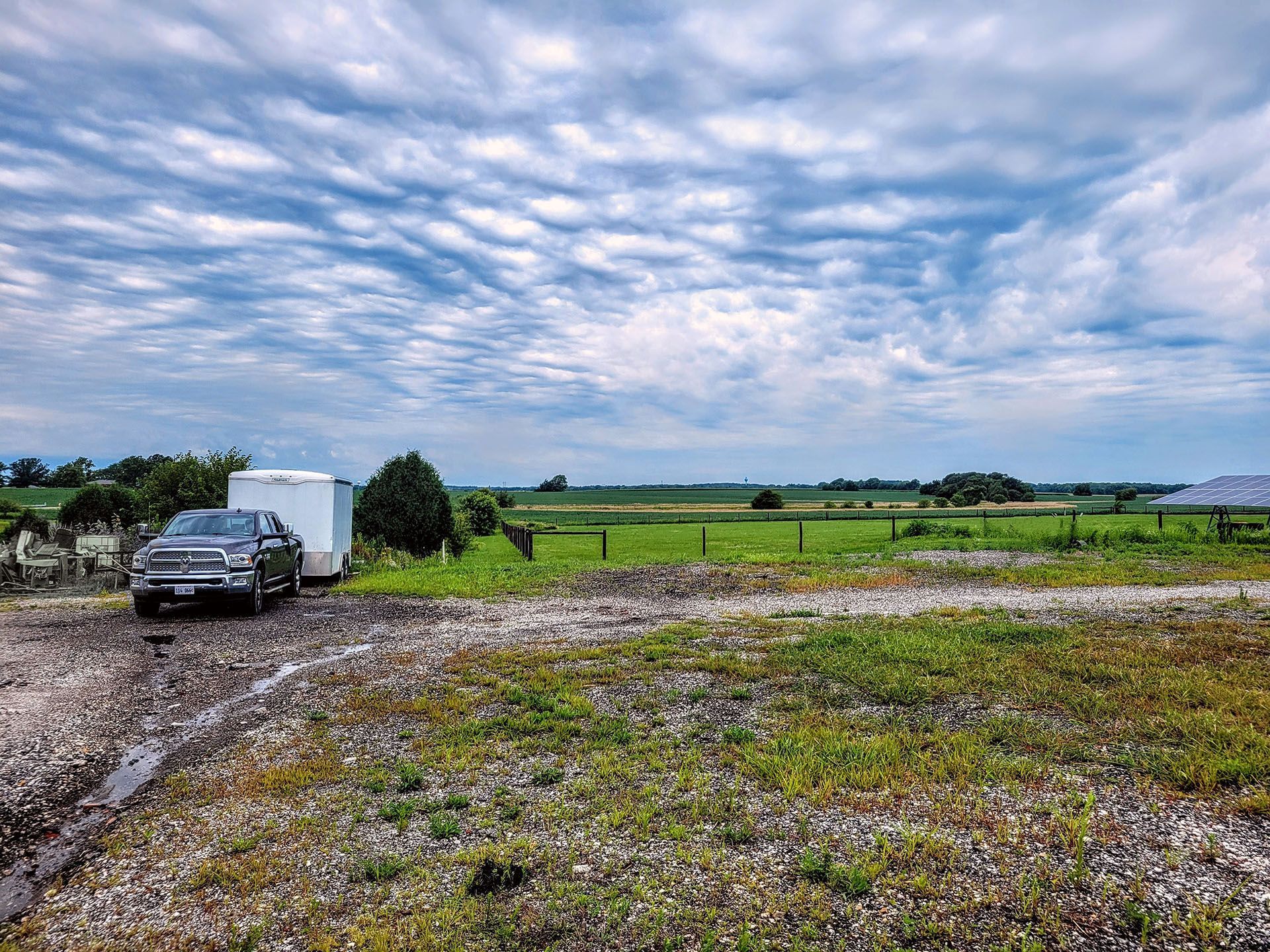 Gray truck and trailer parked on a dirt road, overlooking a green field under a cloudy sky.