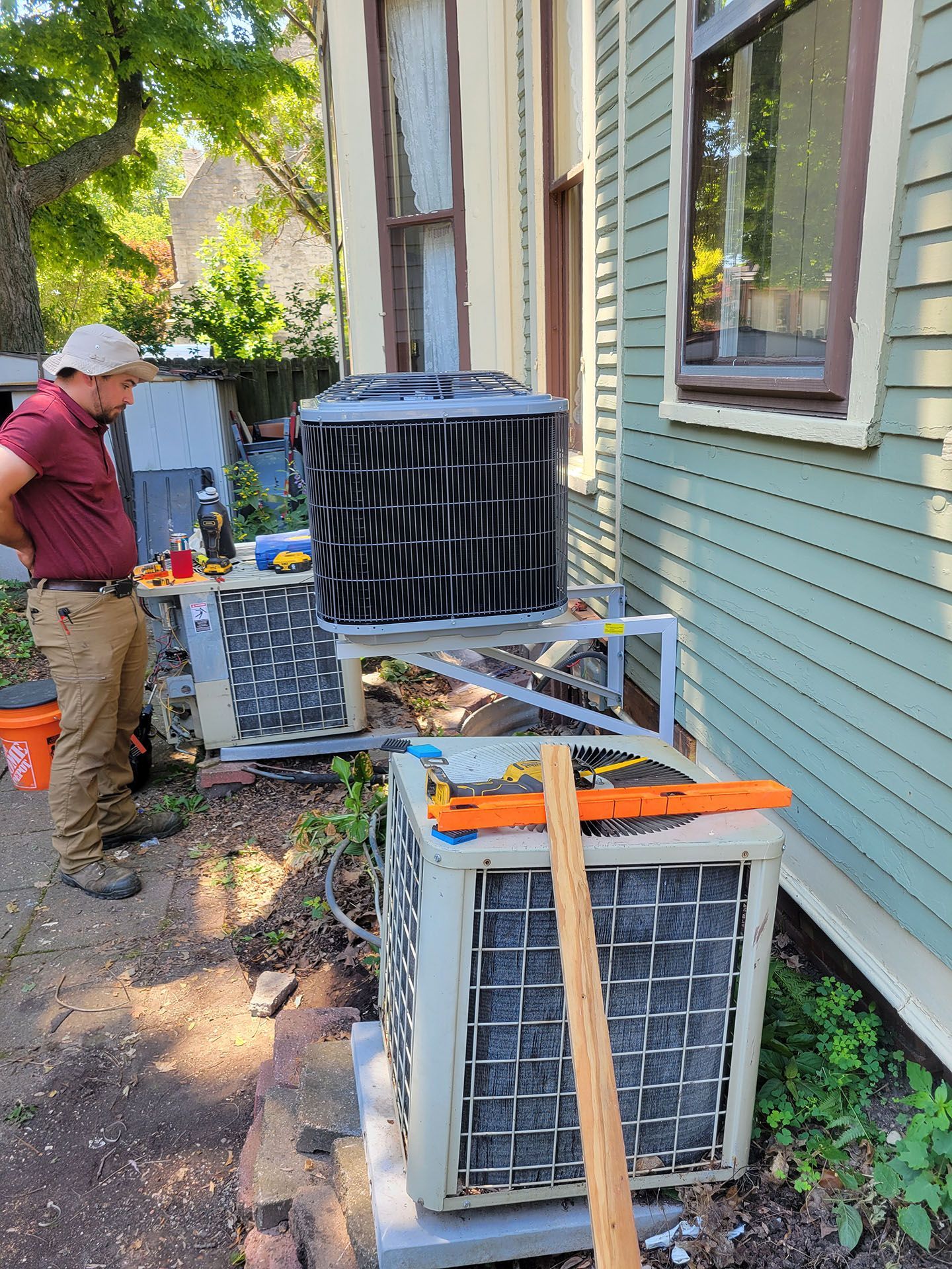 HVAC technician beside two AC units by a house. He is working on the top unit.