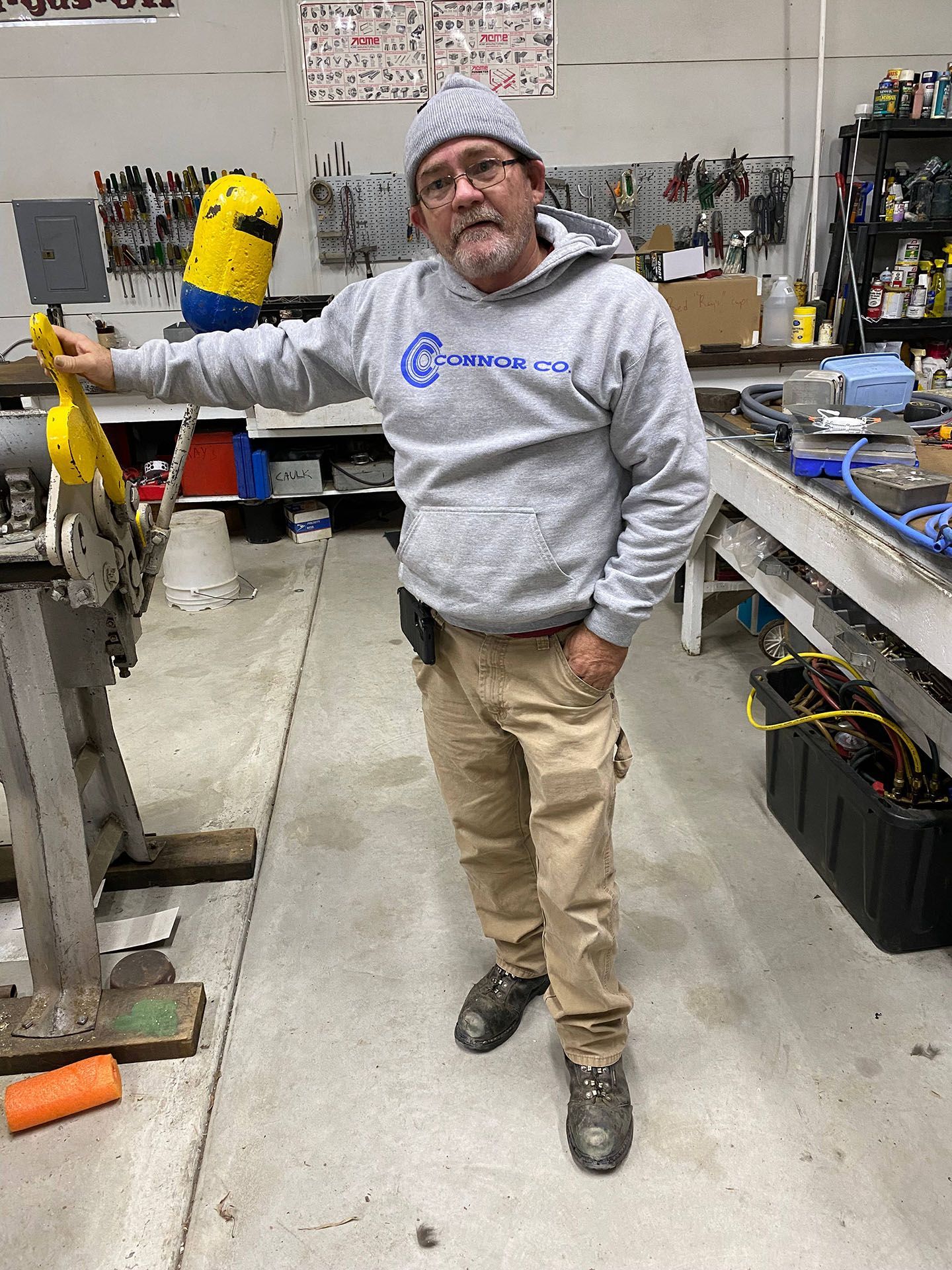Man in workshop wearing a gray hoodie and beanie, holding a tool.