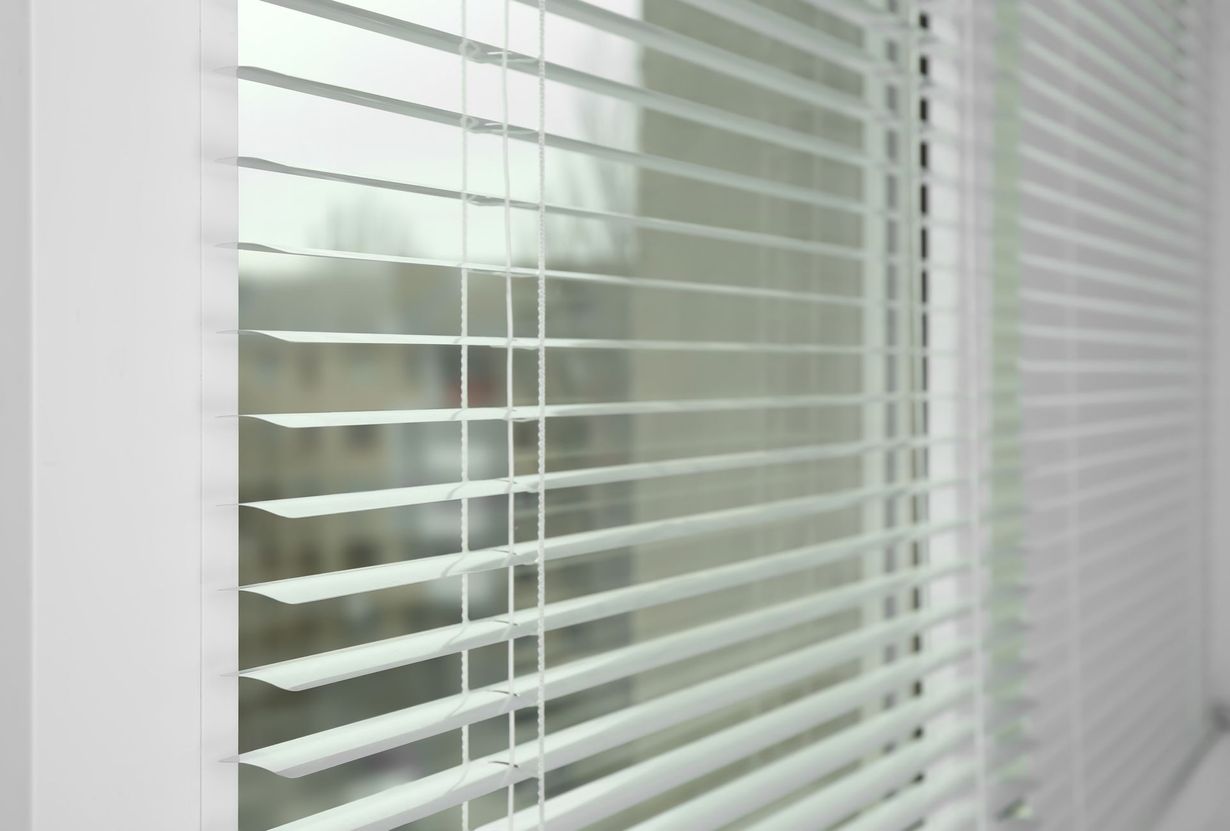 White horizontal window blinds covering a window, with a soft, out-of-focus view of the outdoors visible behind them.