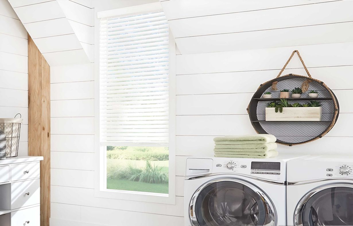 A laundry room with white shiplap walls, a window with white blinds, a round shelf, and two white front-load appliances.