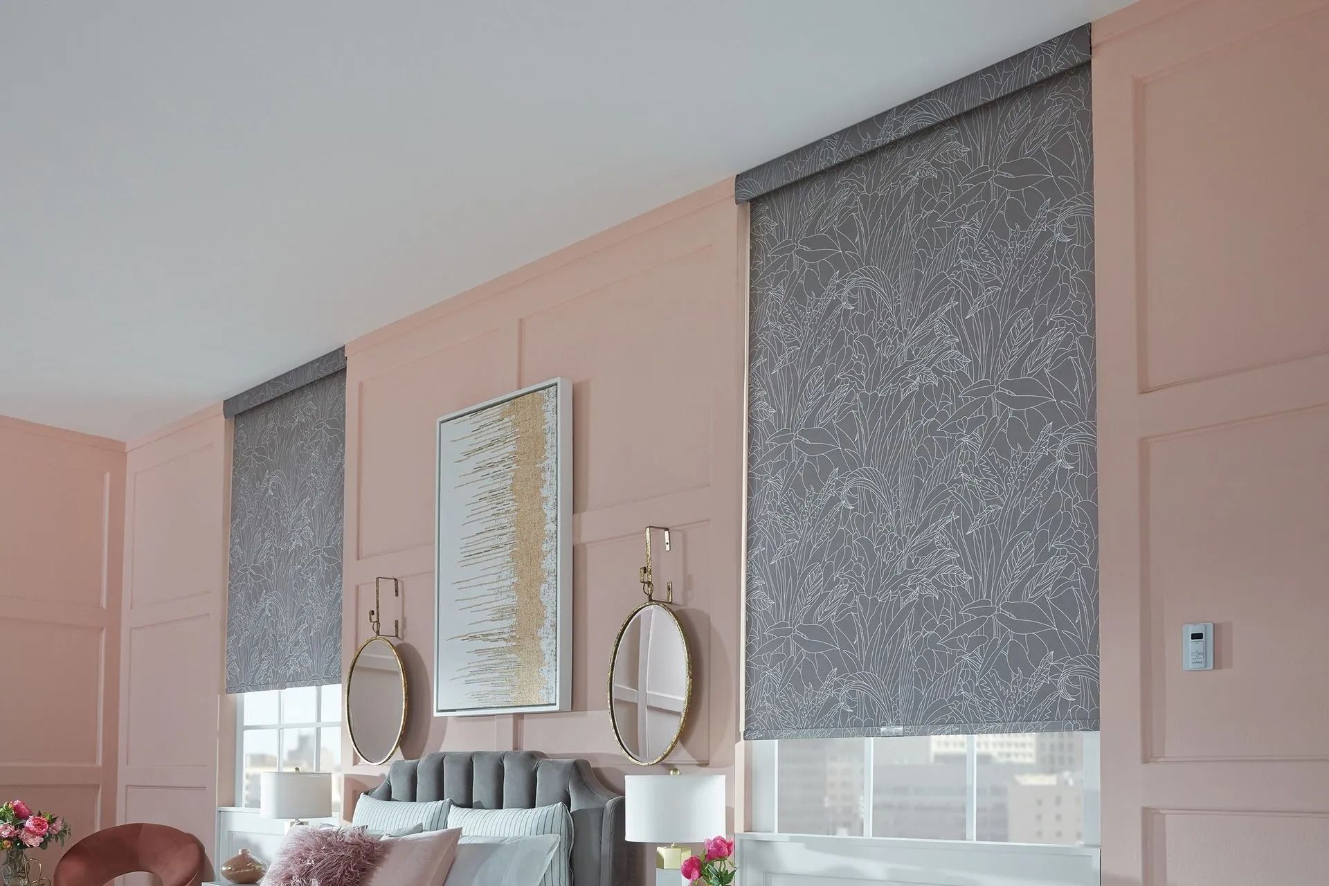 Two gray patterned roller shades hang against a soft pink wall with wainscoting, flanking a central piece of art.