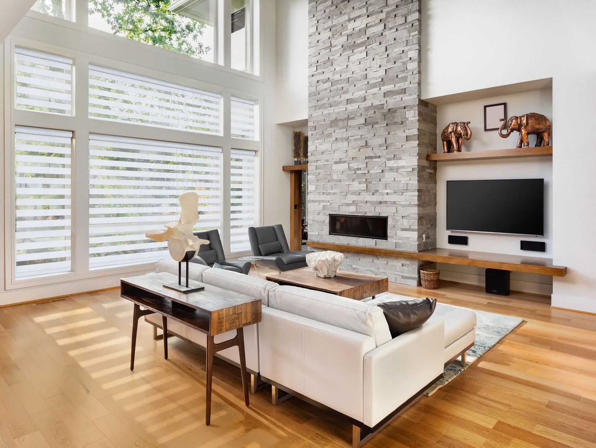 A modern living room with a stone fireplace, light wood floors, a white sofa, and a large window with white blinds.