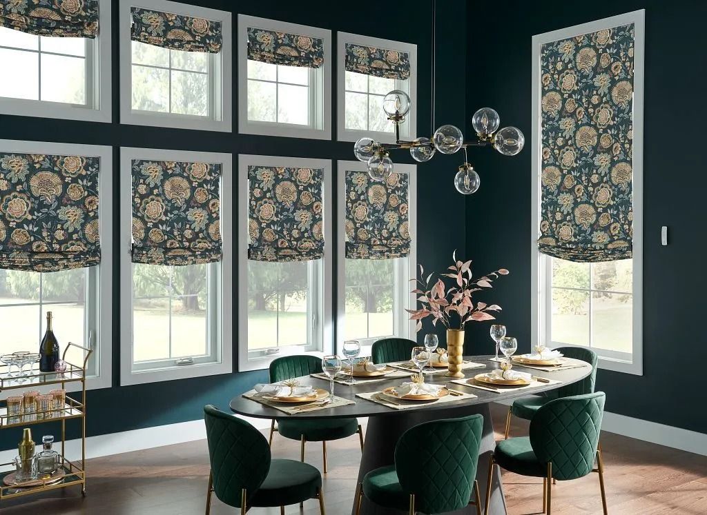 A formal dining room features a dark teal color scheme, a round table with green velvet chairs, and floral Roman shades.