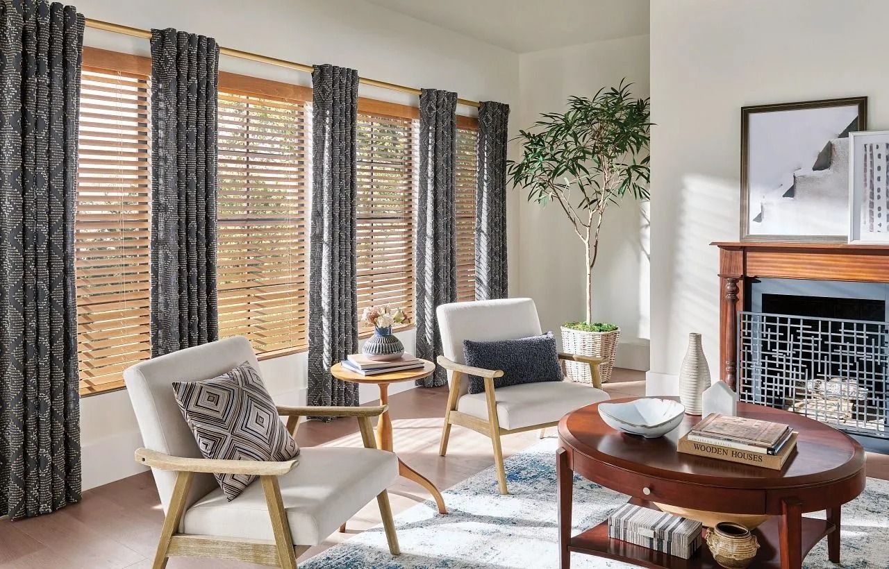 Two neutral-toned armchairs face a wooden coffee table in front of a window with patterned curtains and wood blinds.