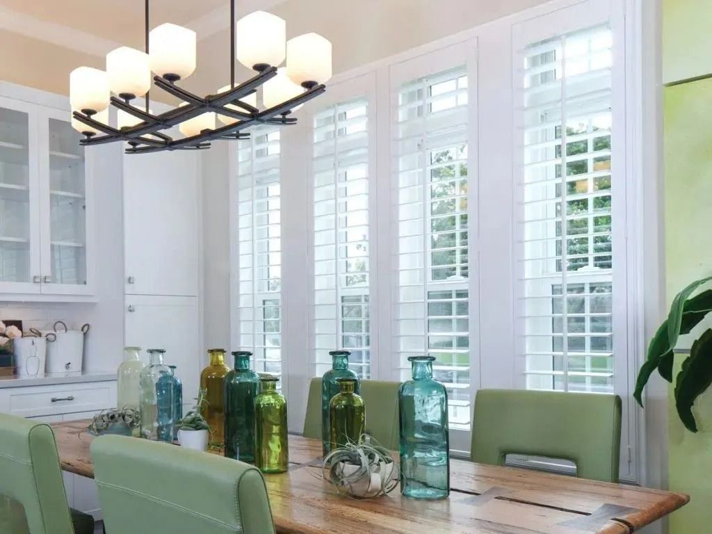 A dining room featuring a wooden table with colorful glass vases, green chairs, a modern chandelier, and shuttered windows.