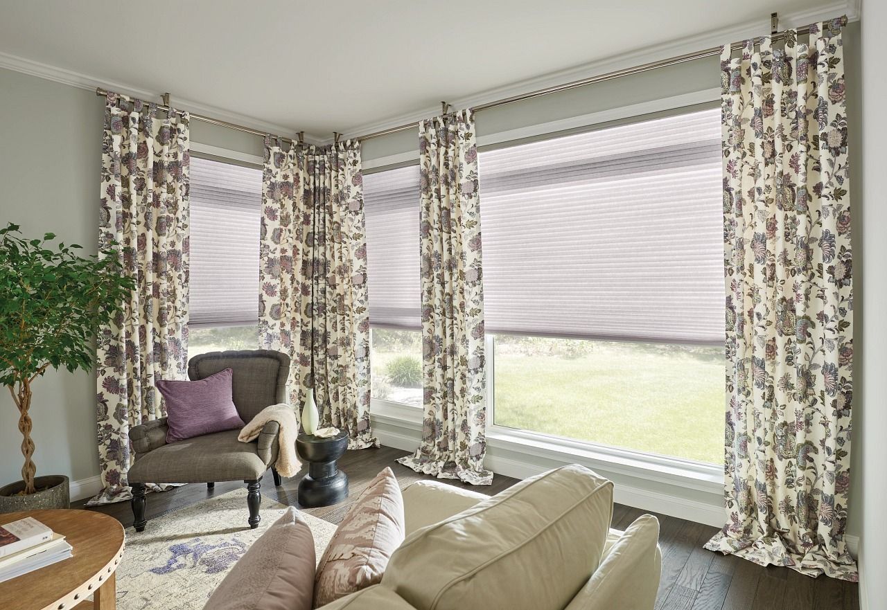 Living room with patterned curtains, cellular shades, a dark armchair with a purple pillow, and a beige sofa.