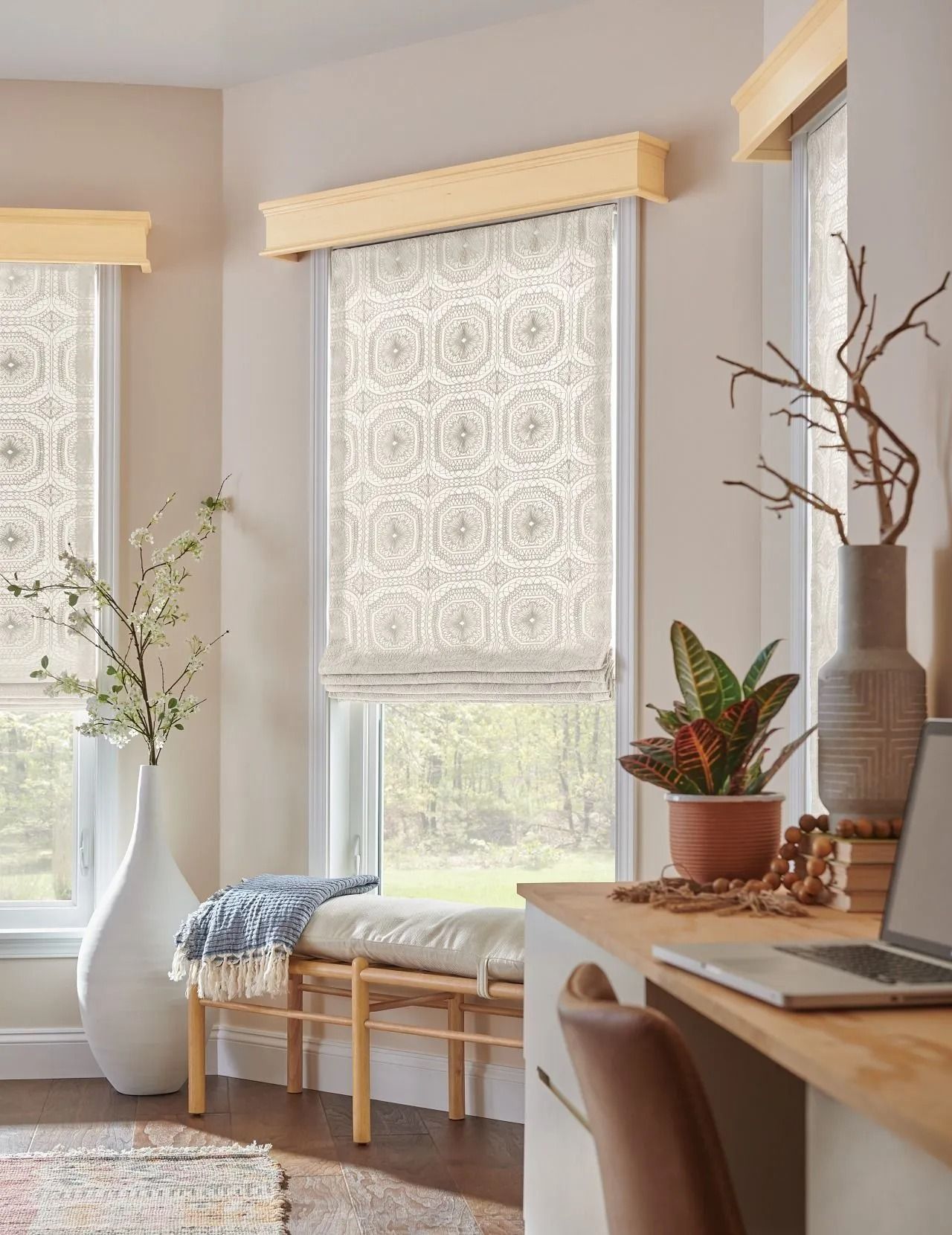 Three windows with patterned Roman shades and wooden valances, adjacent to a desk, a bench, and indoor plants.