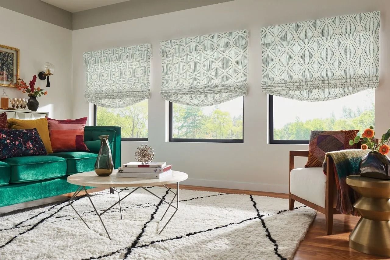 A living room with three windows featuring patterned Roman shades, a green velvet sofa, white armchair, and shag rug.