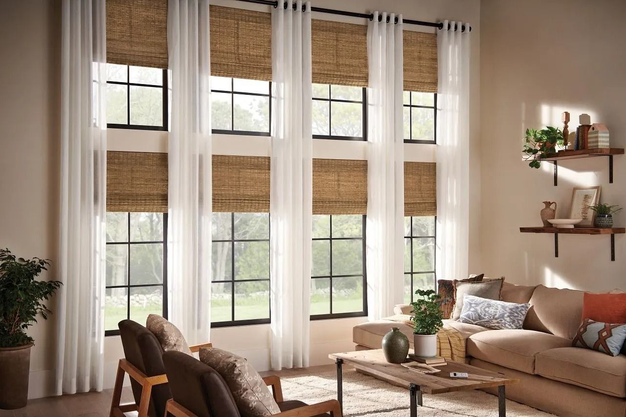 Living room with large windows, light curtains, woven shades, a tan sofa, and wall-mounted shelves with decor.