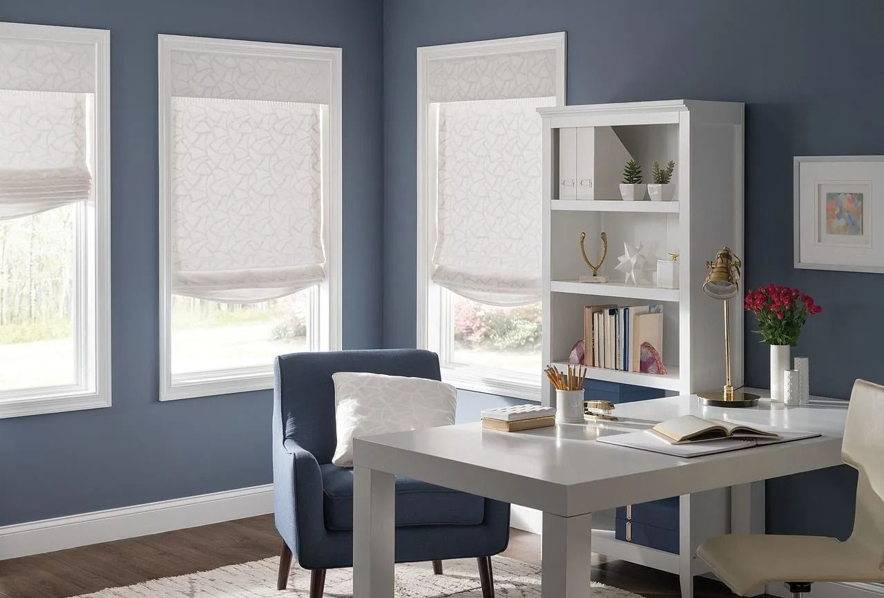 A home office with dark blue walls, three windows with white Roman shades, a white desk, a blue armchair, and a bookshelf.
