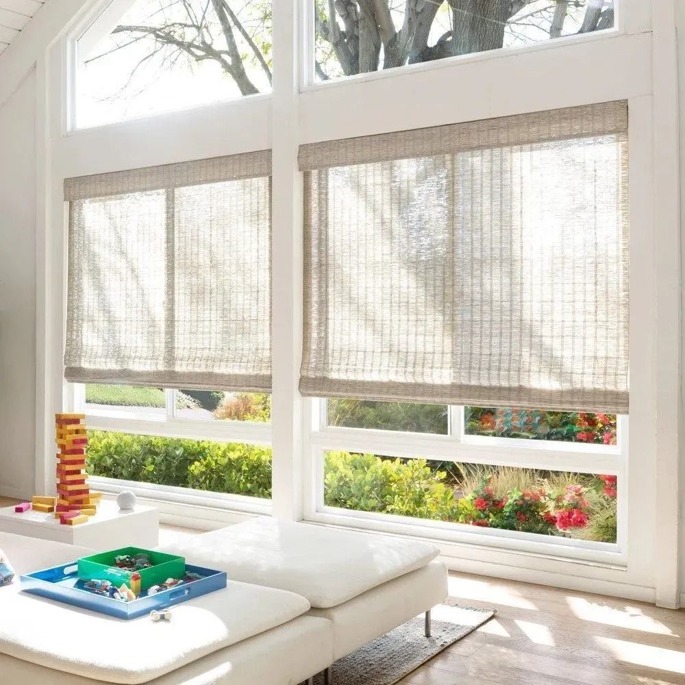 Two large windows covered with light-colored woven shades, overlooking a sunny outdoor garden scene.