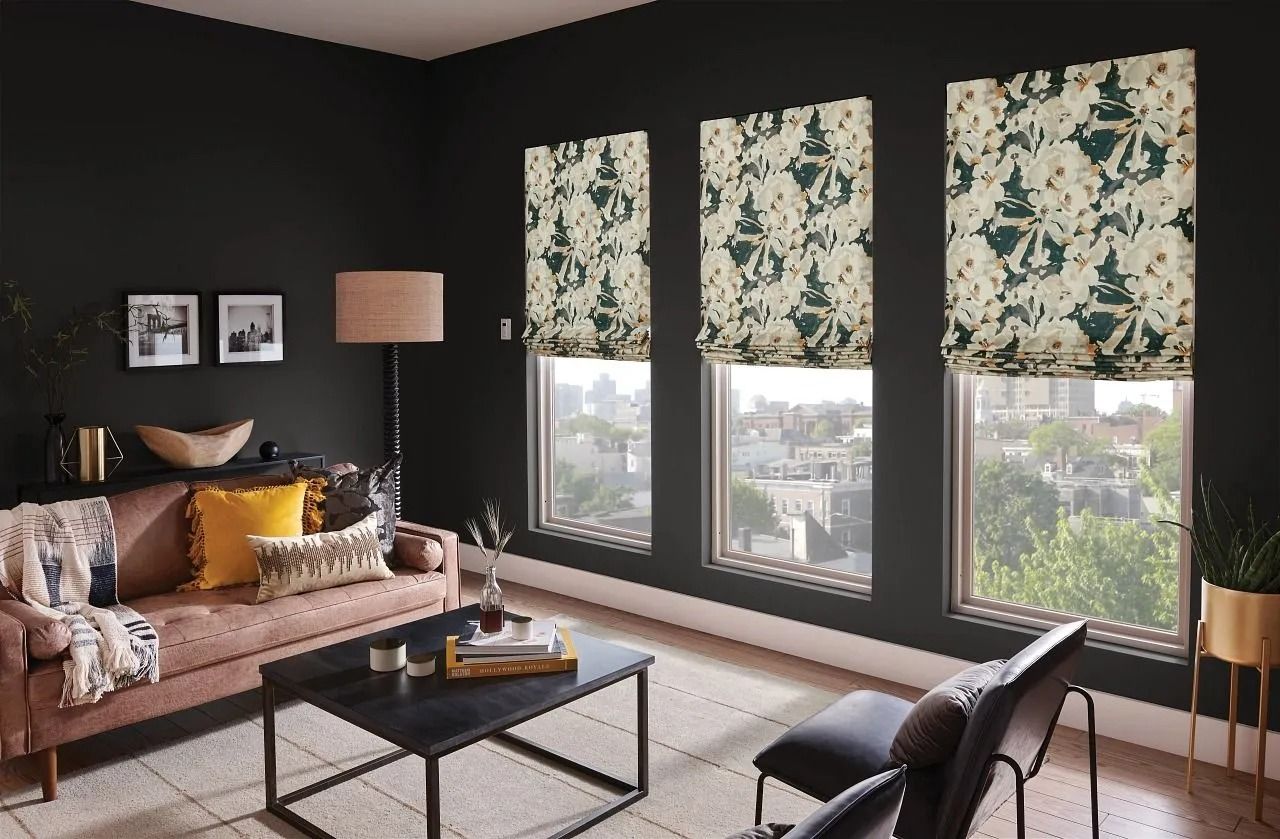 A modern living room with a pink sofa, dark walls, and three windows adorned with patterned floral Roman shades.