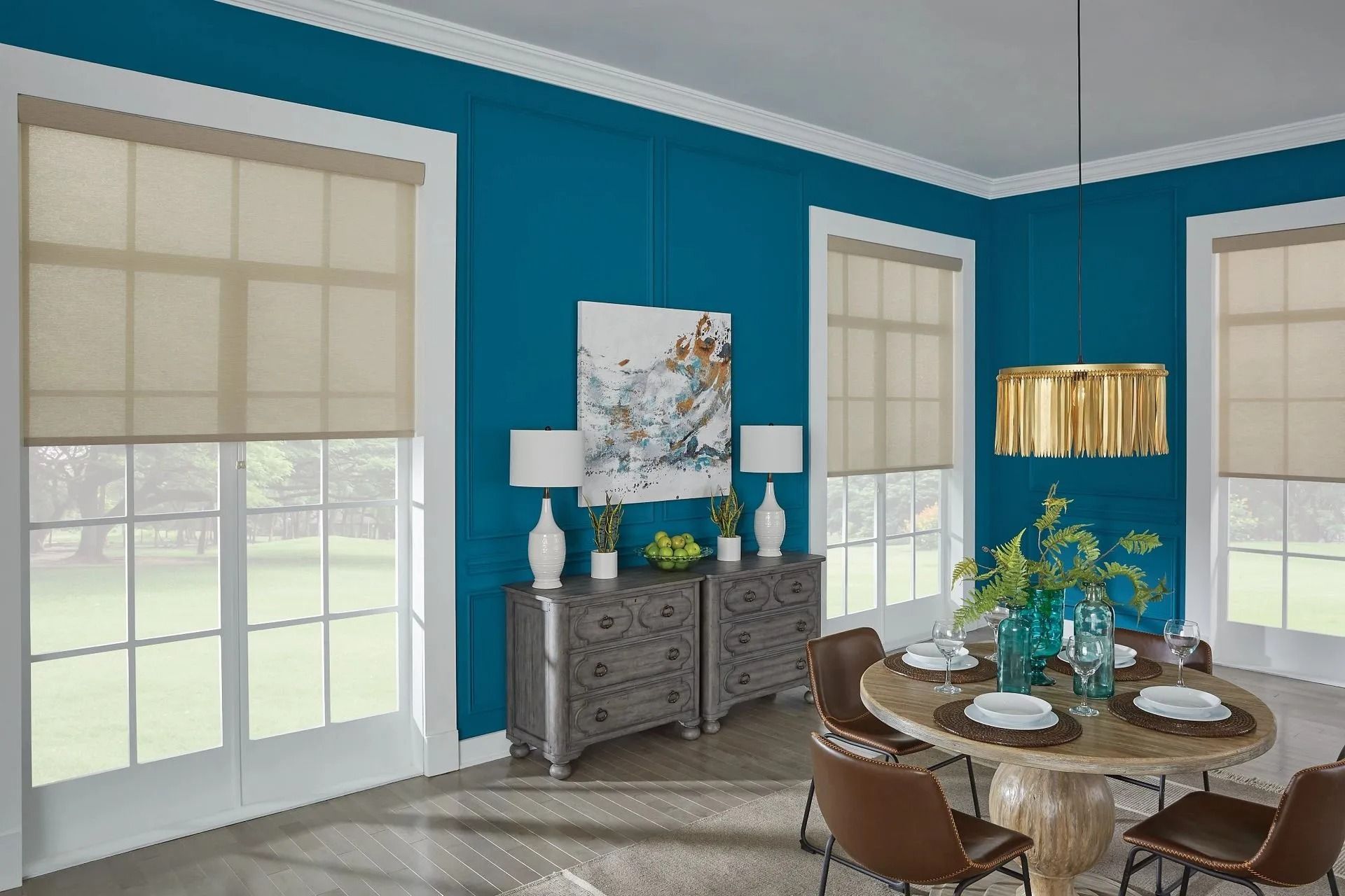 A dining room with teal walls, beige roller shades on large windows, a wooden table with chairs, and a grey sideboard.
