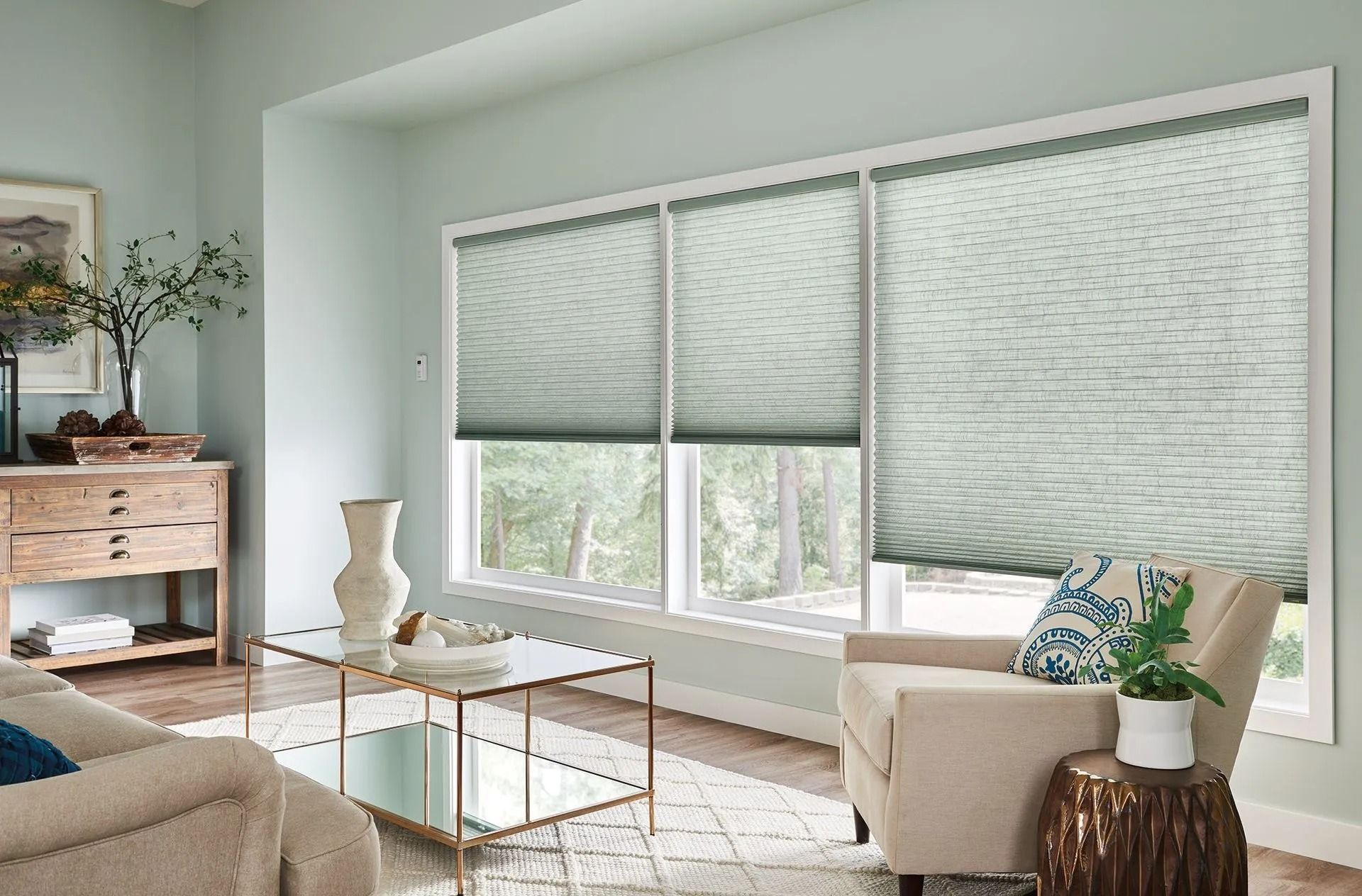 A sunlit living room featuring three large windows covered by light-filtering honeycomb shades with a beige armchair.