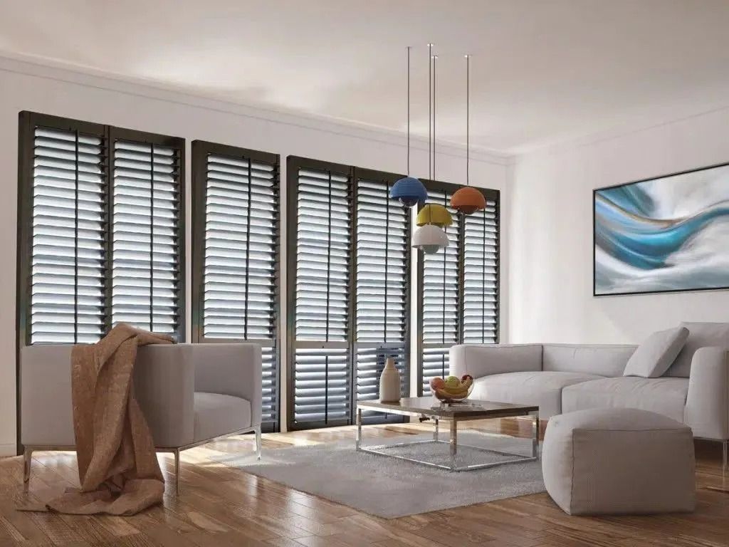 A modern living room with large windows, dark shutters, grey sofa, armchair, coffee table, and colorful pendant lights.