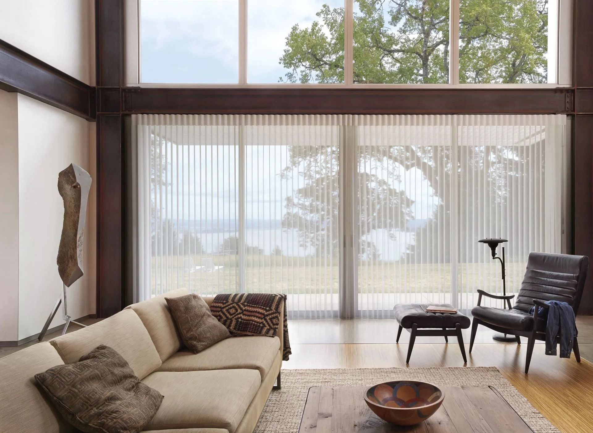 A modern living room with a beige couch, a leather armchair, and large windows with sheer floor-to-ceiling blinds.