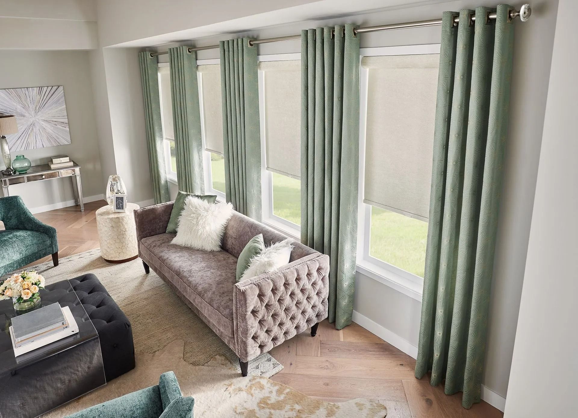 A living room with a tufted sofa, sage green curtains over roller shades, and a dark ottoman on a patterned rug.