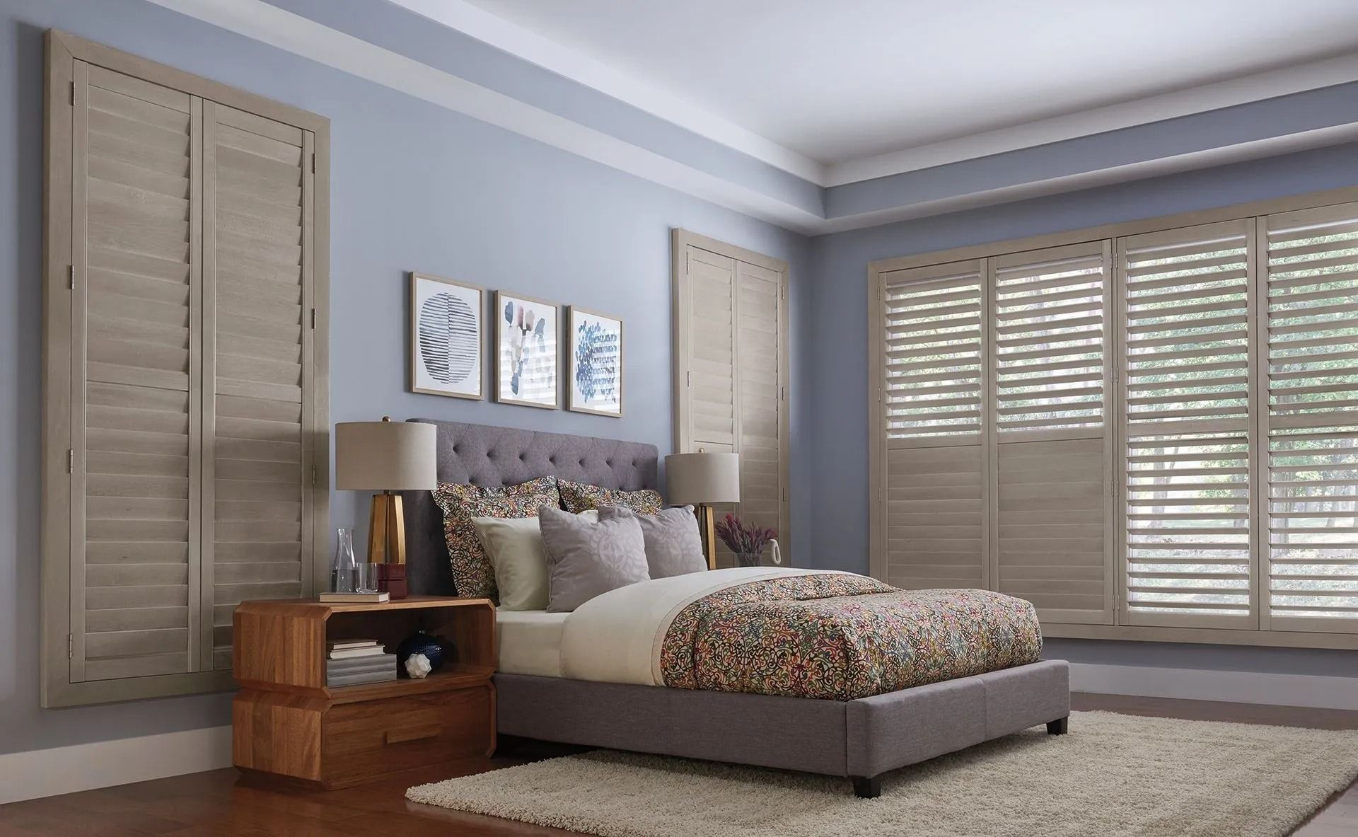 A bedroom with a tufted gray bed, three wall prints, a wood nightstand, and light-toned plantation shutters on the walls.