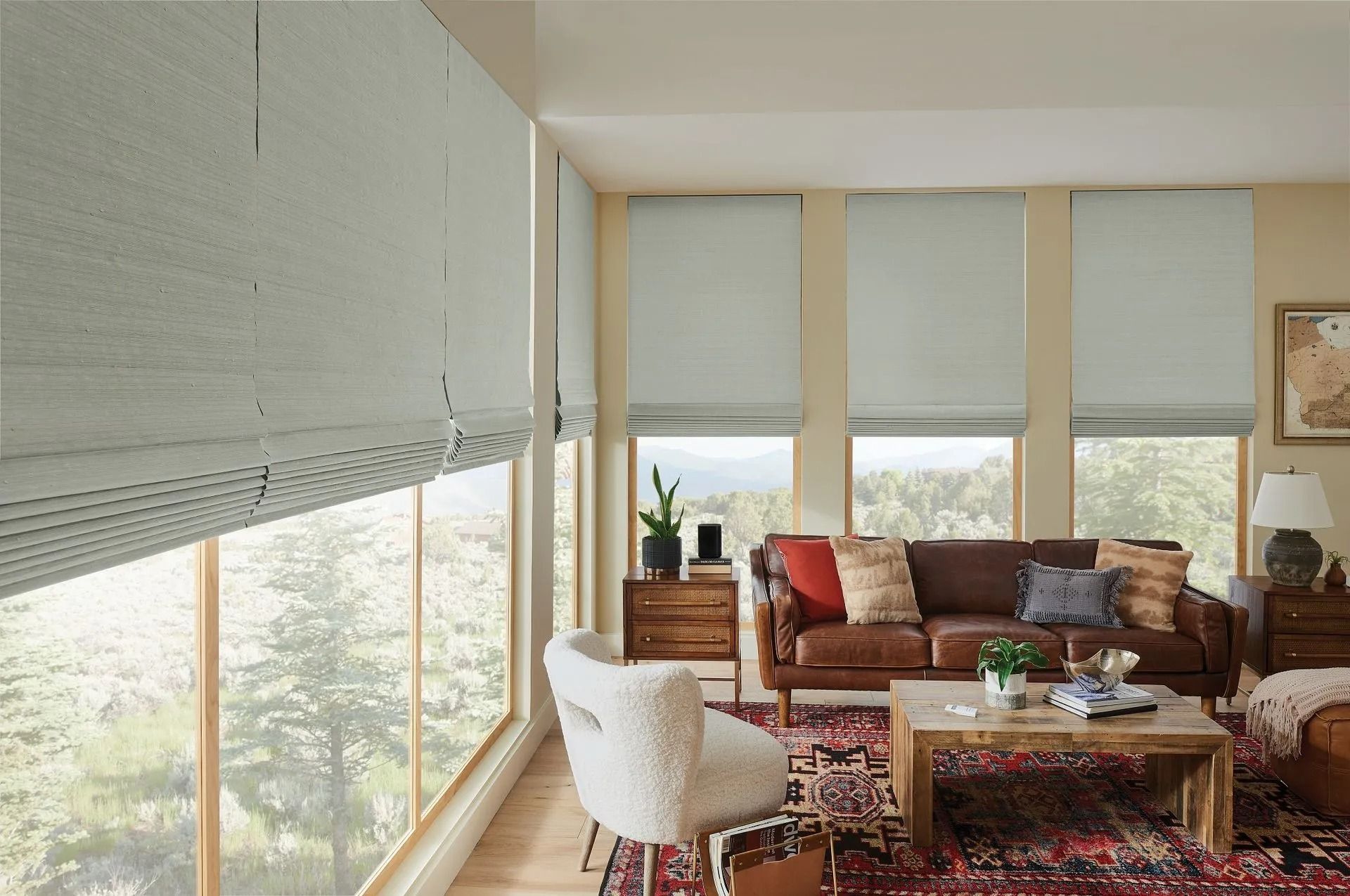 A cozy living room with large windows, light-colored Roman shades, a brown leather sofa, and patterned area rug.
