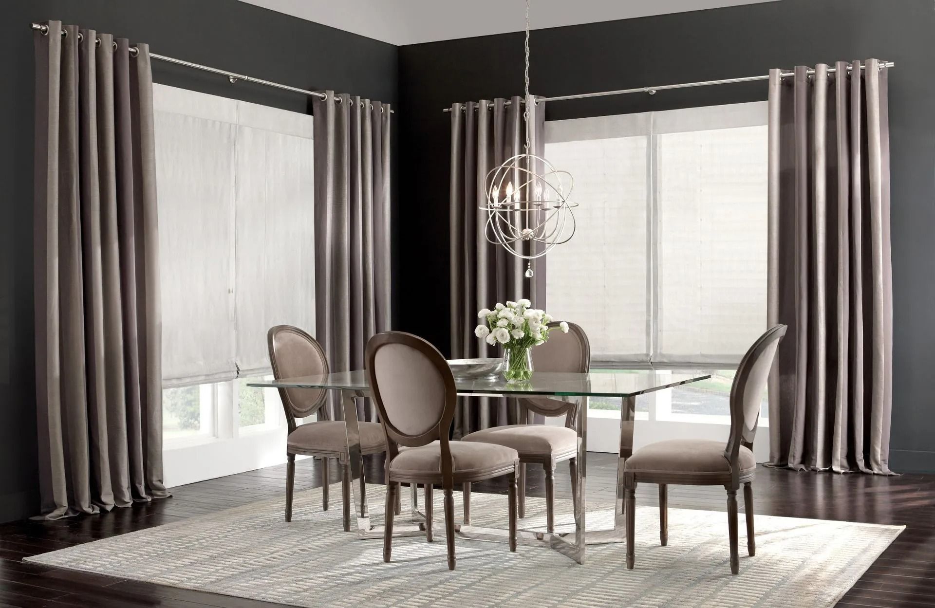 Dining room with a glass table, four upholstered chairs, taupe curtains, and white window shades against dark walls.