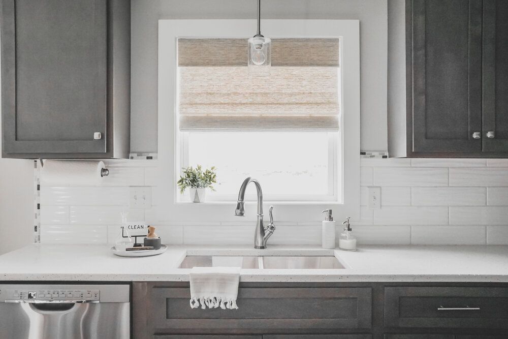 A modern kitchen sink area with dark cabinetry, white subway tile backsplash, a window with a woven shade, and faucet.