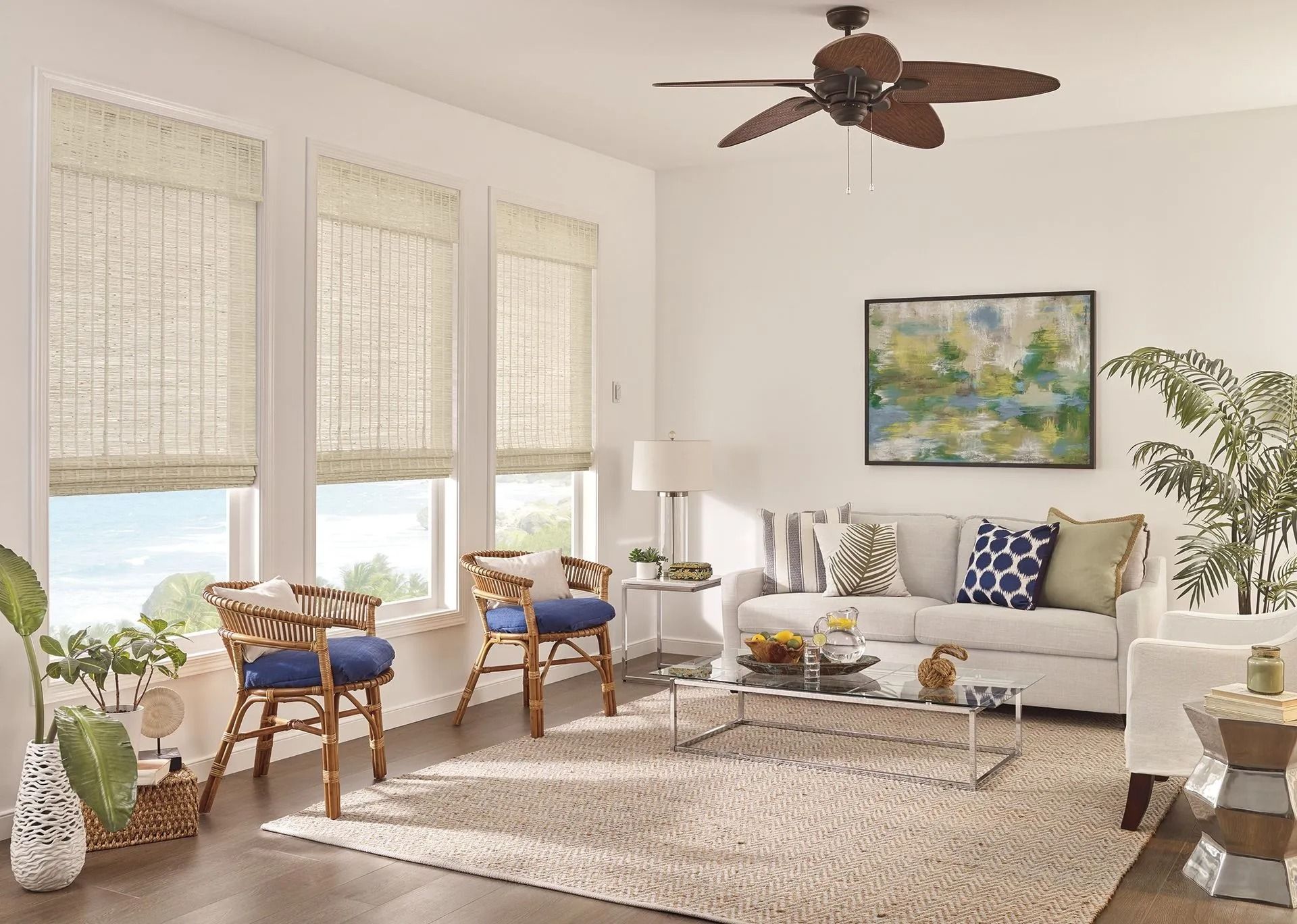 Living room with a beige sofa, two rattan chairs with blue cushions, large windows with bamboo shades, and a ceiling fan.
