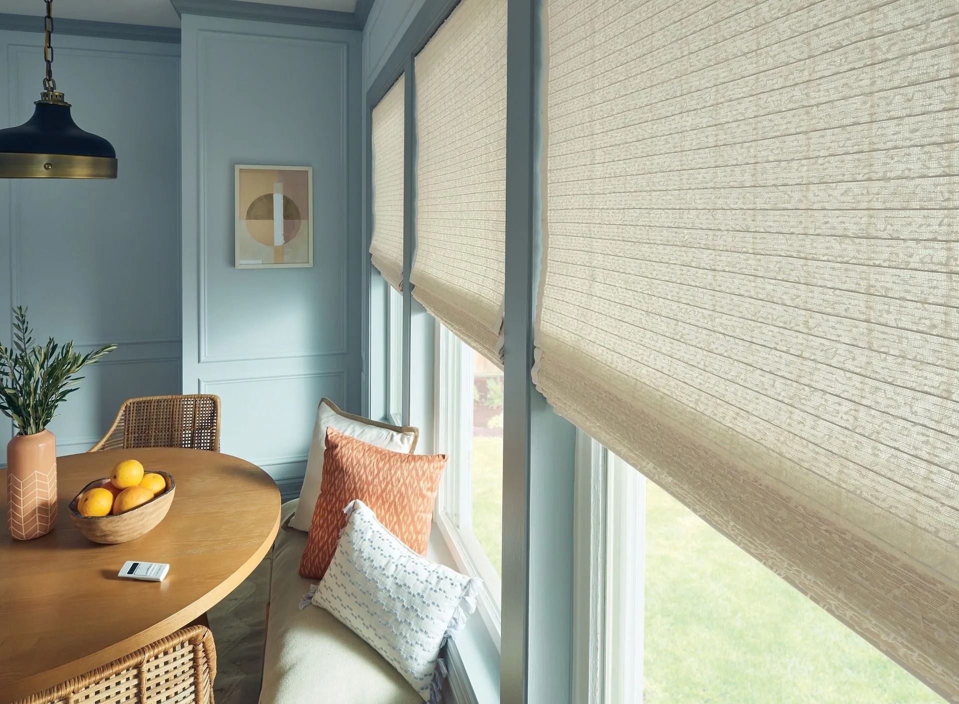 A dining area with a round wooden table, a bowl of lemons, textured window shades, and a cushioned bench with pillows.