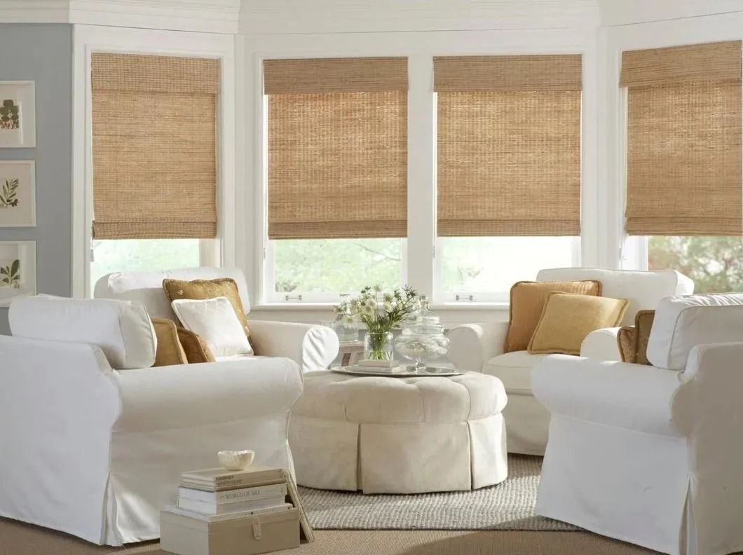 A living room area with white armchairs arranged around a round, upholstered ottoman in front of a bay window.