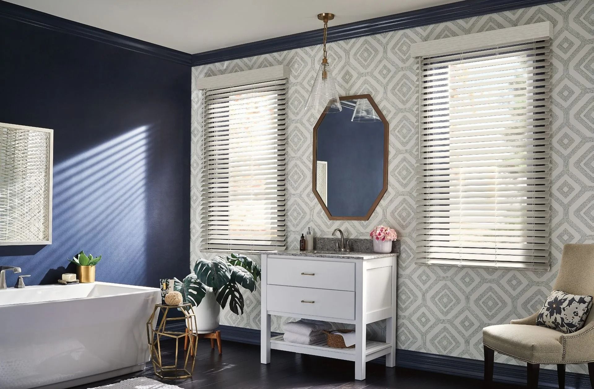 A bathroom with a white vanity, a mirror, two windows with blinds, a white bathtub, and patterned wallpaper.