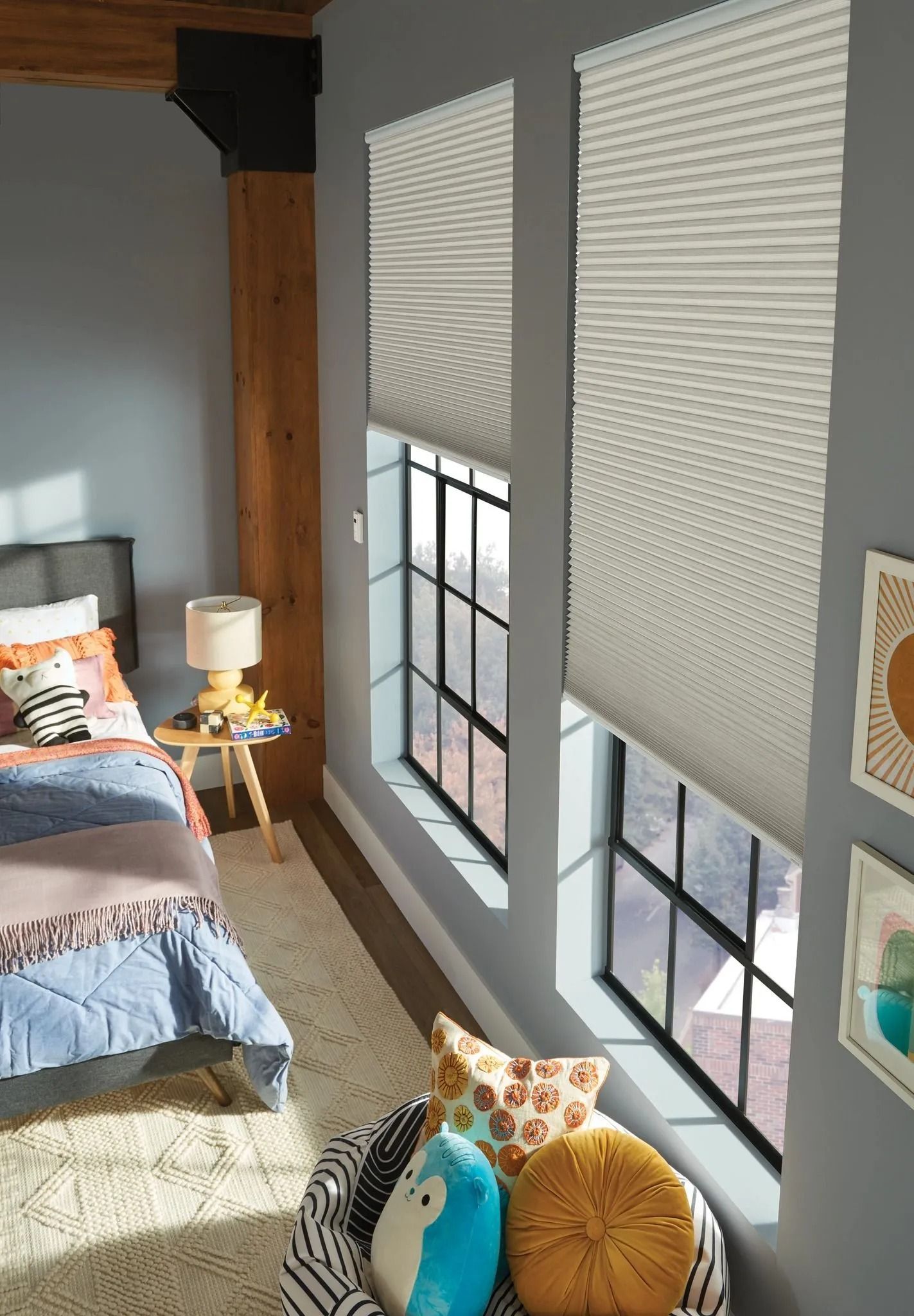 A bedroom with grey walls features windows covered by pleated shades, a bed with colorful bedding, and a textured rug.