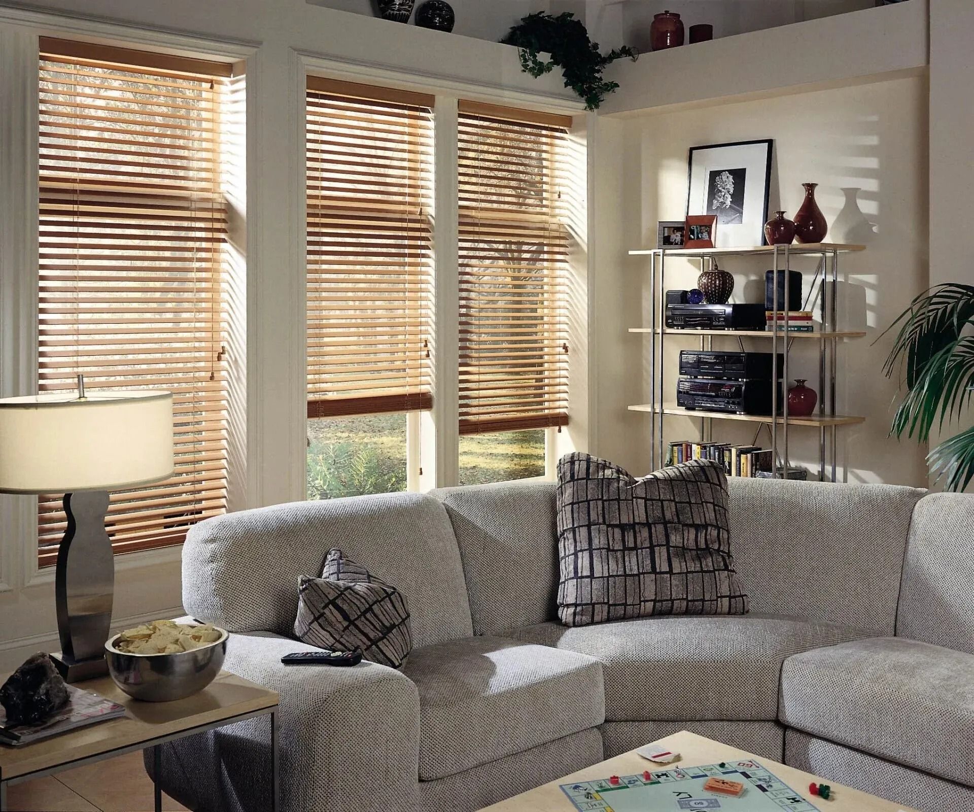 A light-colored sectional sofa faces a shelving unit and three large windows with wooden blinds in a living room.
