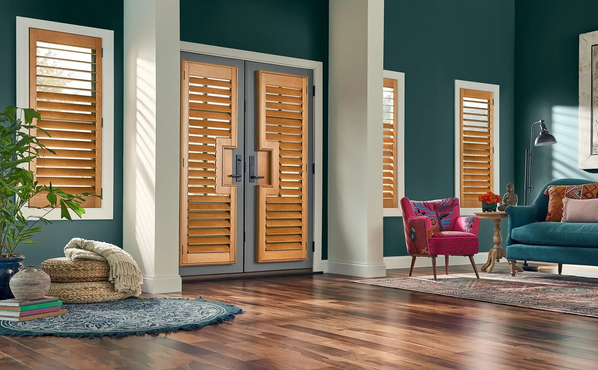A teal living room with wooden louvered shutters on a double door and two windows, featuring a patterned chair and sofa.