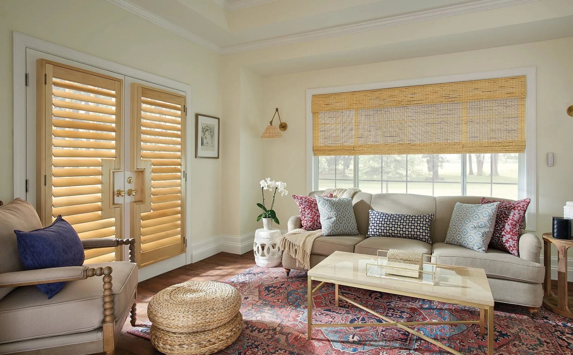 A light-filled living room with a beige sofa, patterned rug, wooden shutters on French doors, and woven blinds on a window.