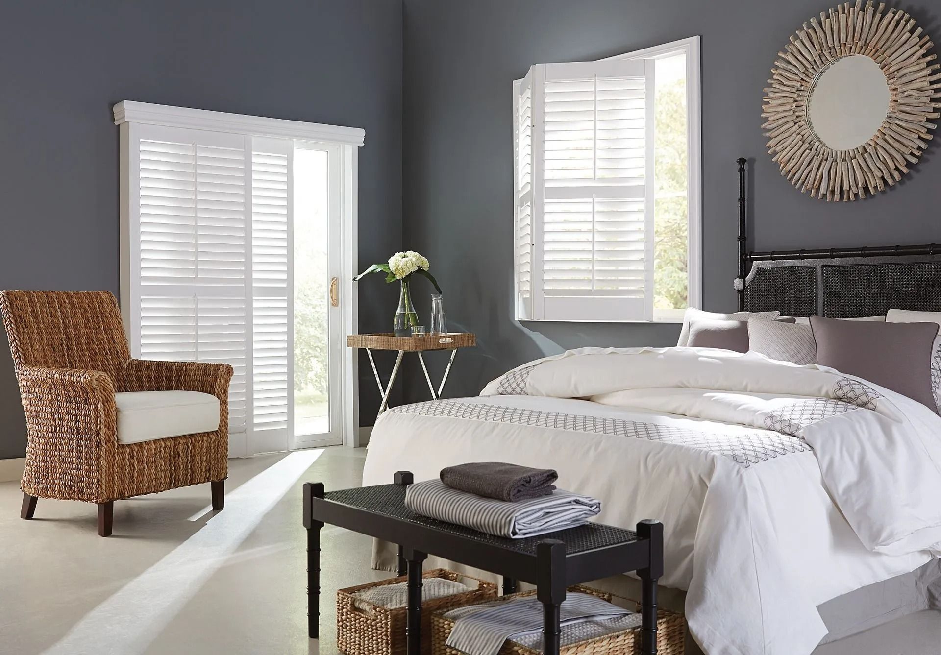 A bedroom with dark gray walls, a bed with white bedding, a wicker chair, white window shutters, and a round wall mirror.