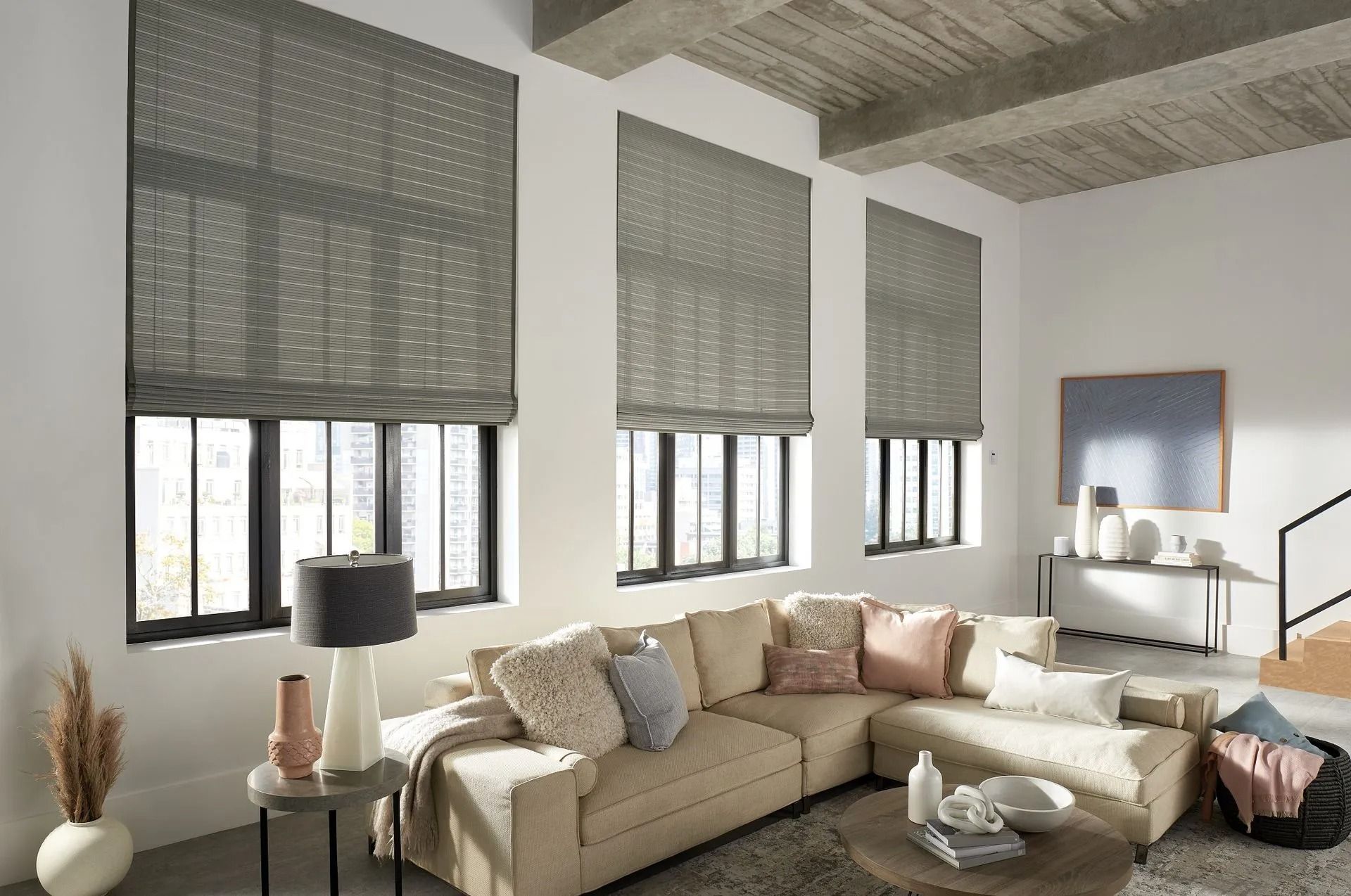 A modern living room with beige sectional sofa, grey window shades, and neutral decor under a wood-beamed ceiling.