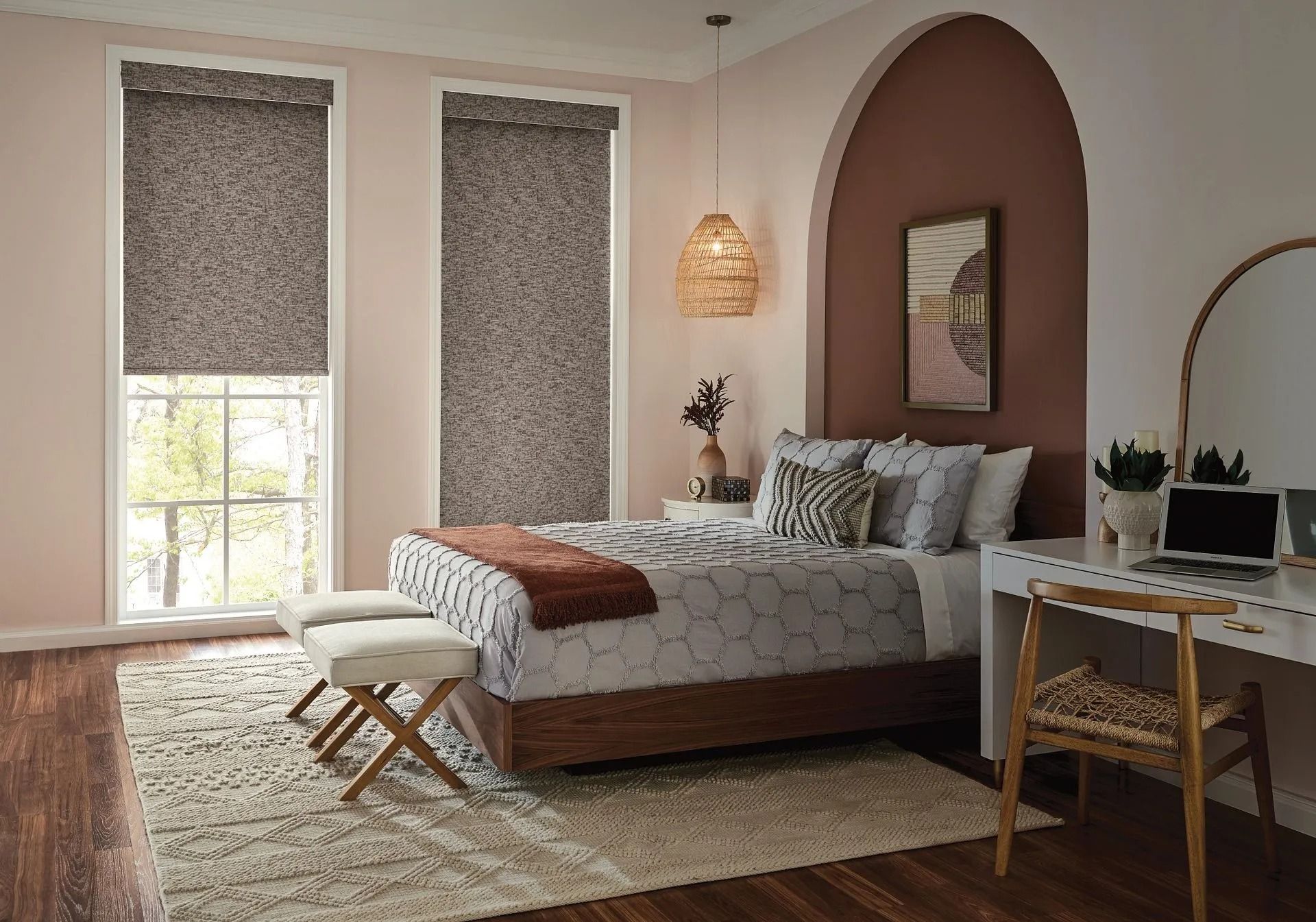 A bedroom with two patterned roller shades, a bed with gray bedding, a rustic desk, and a textured rug on a wood floor.