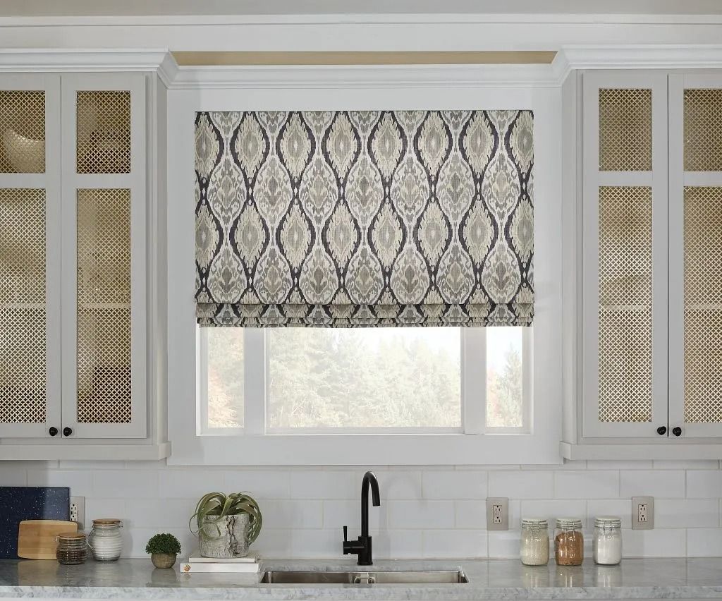 A kitchen window with a patterned grey and white Roman shade, flanked by white cabinets with textured glass panels.