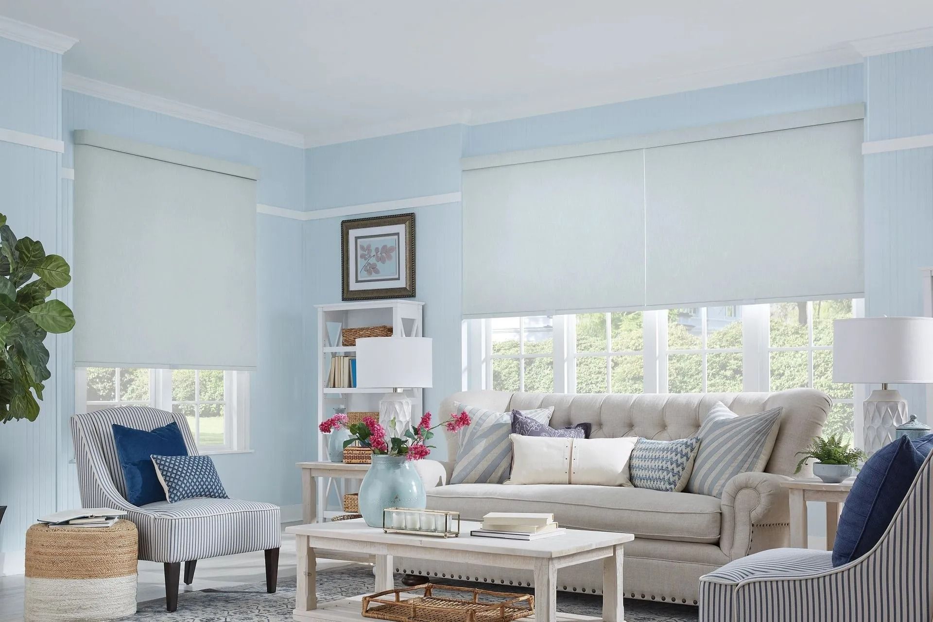 A light blue living room with a beige tufted sofa, striped armchairs, a wooden coffee table, and white roller shades.