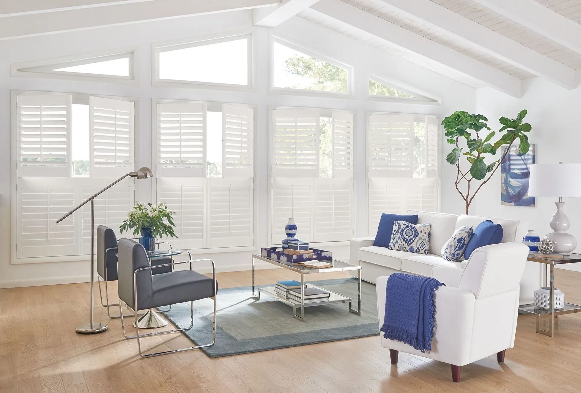 A sunlit living room with white plantation shutters, a grey armchair, a white sofa with blue accents, and a coffee table.