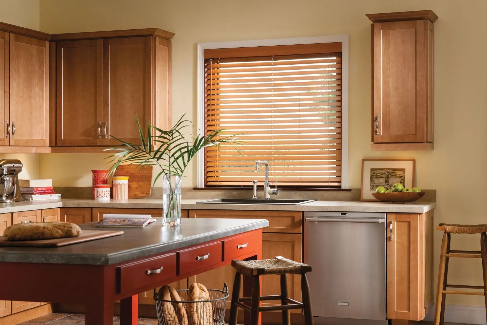 Kitchen interior featuring light wooden cabinets, a dark red island with a stone top, a stainless dishwasher, and blinds.