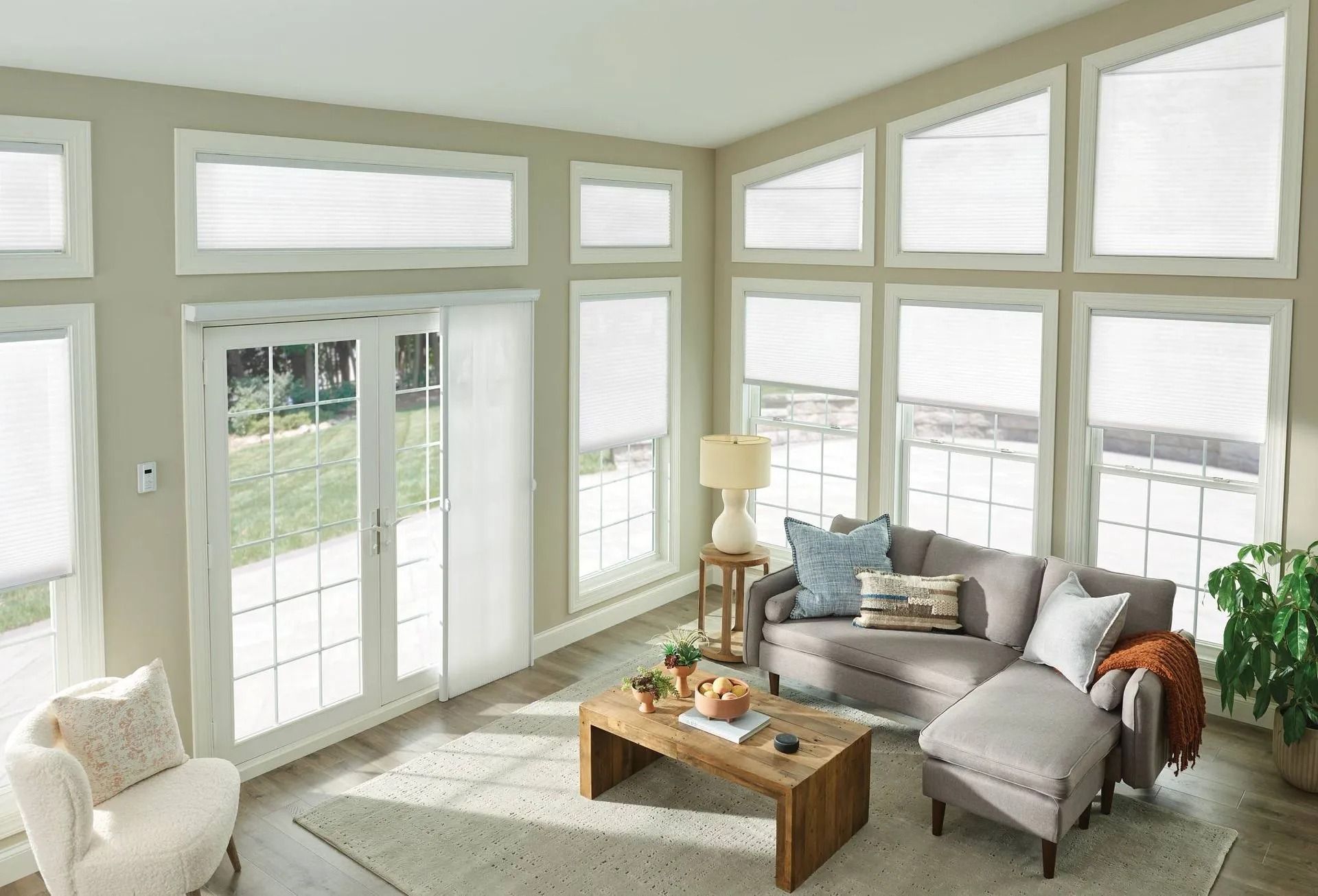 A living room with light walls, a grey sectional sofa, wood coffee table, and large windows with white pleated shades.